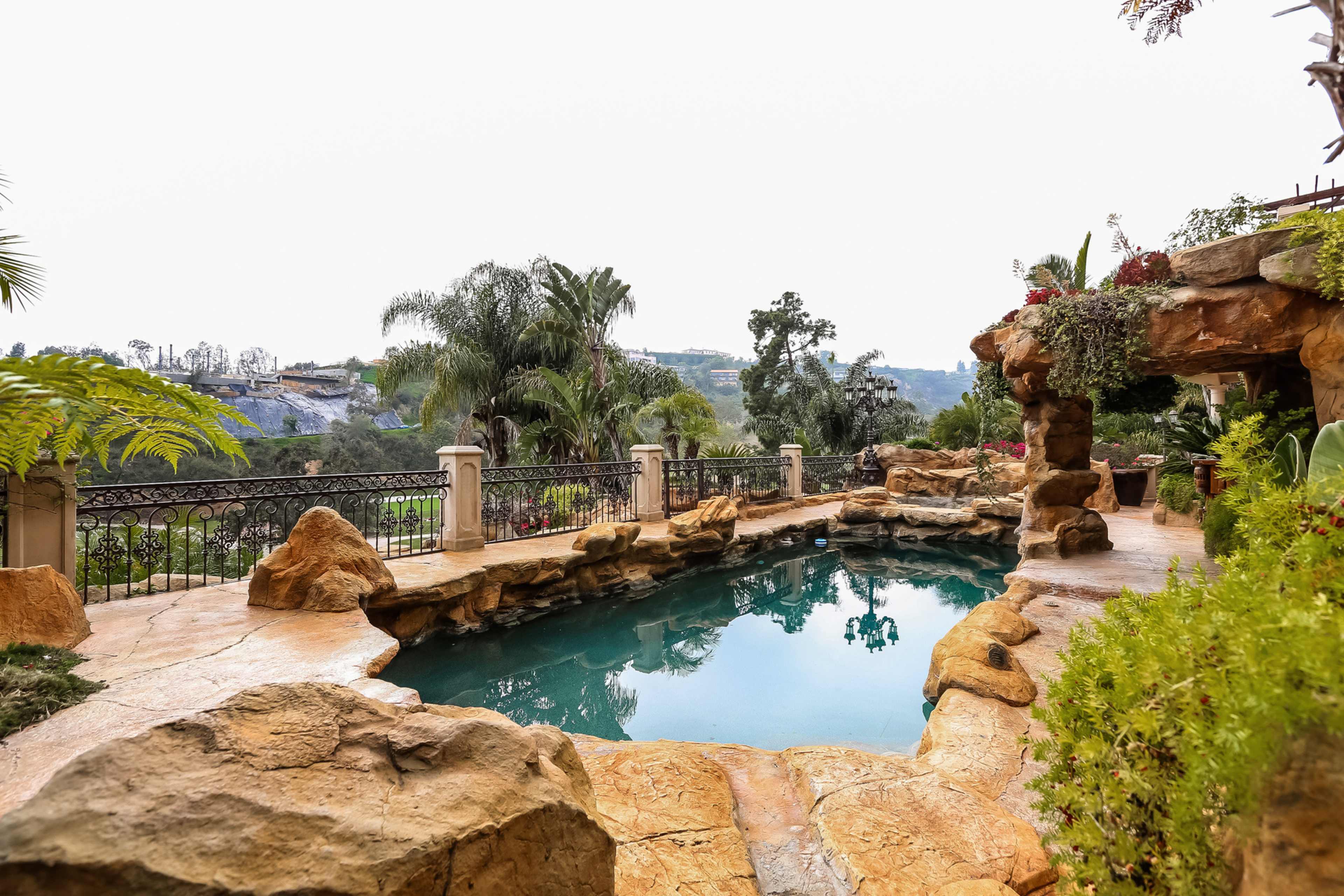 A stone-lined swimming pool is surrounded by lush greenery and palm trees, with a scenic hillside visible in the background.