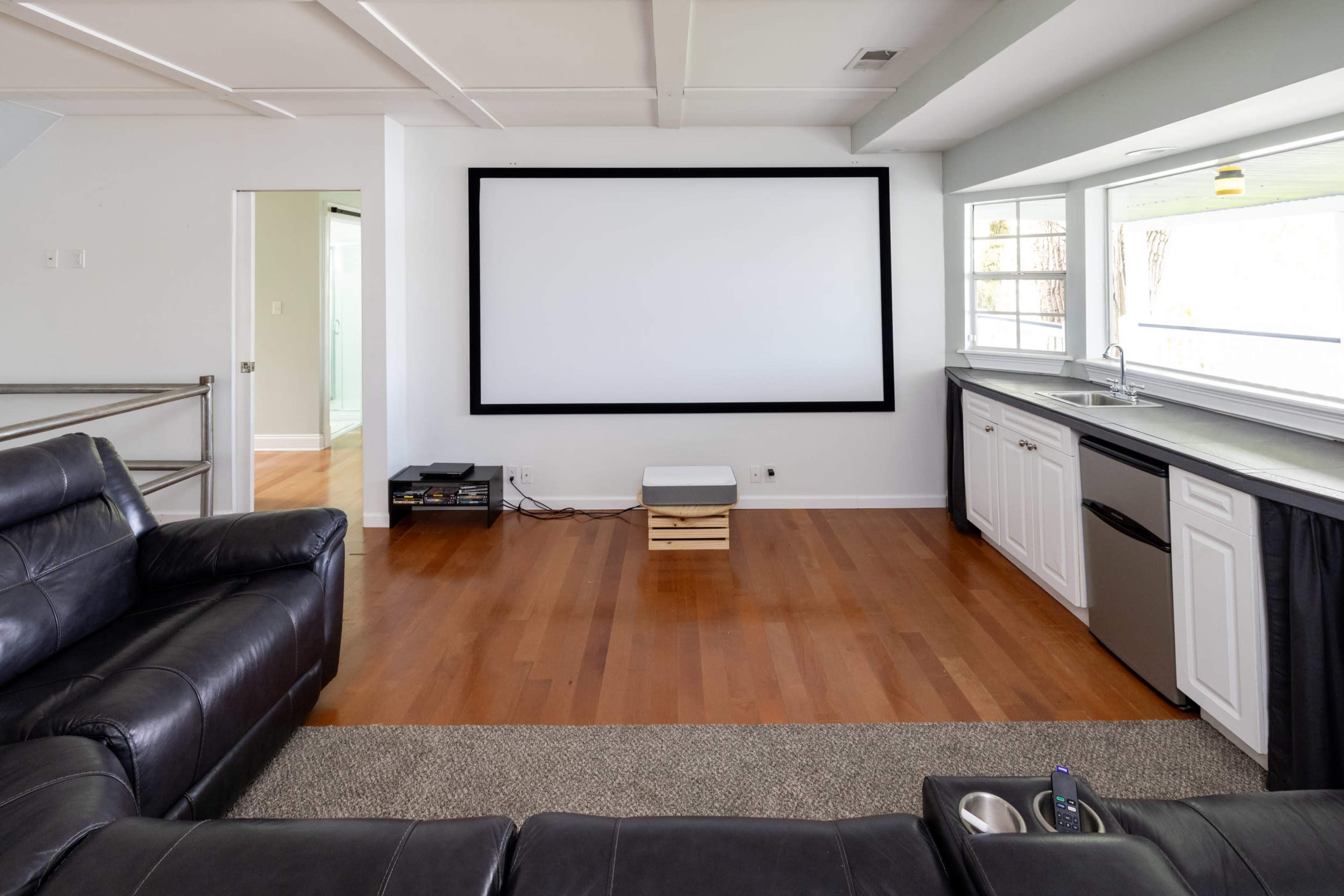 The image shows a spacious living room featuring a large projection screen on one wall, a sectional black sofa, and a kitchenette area with a sink and refrigerator.