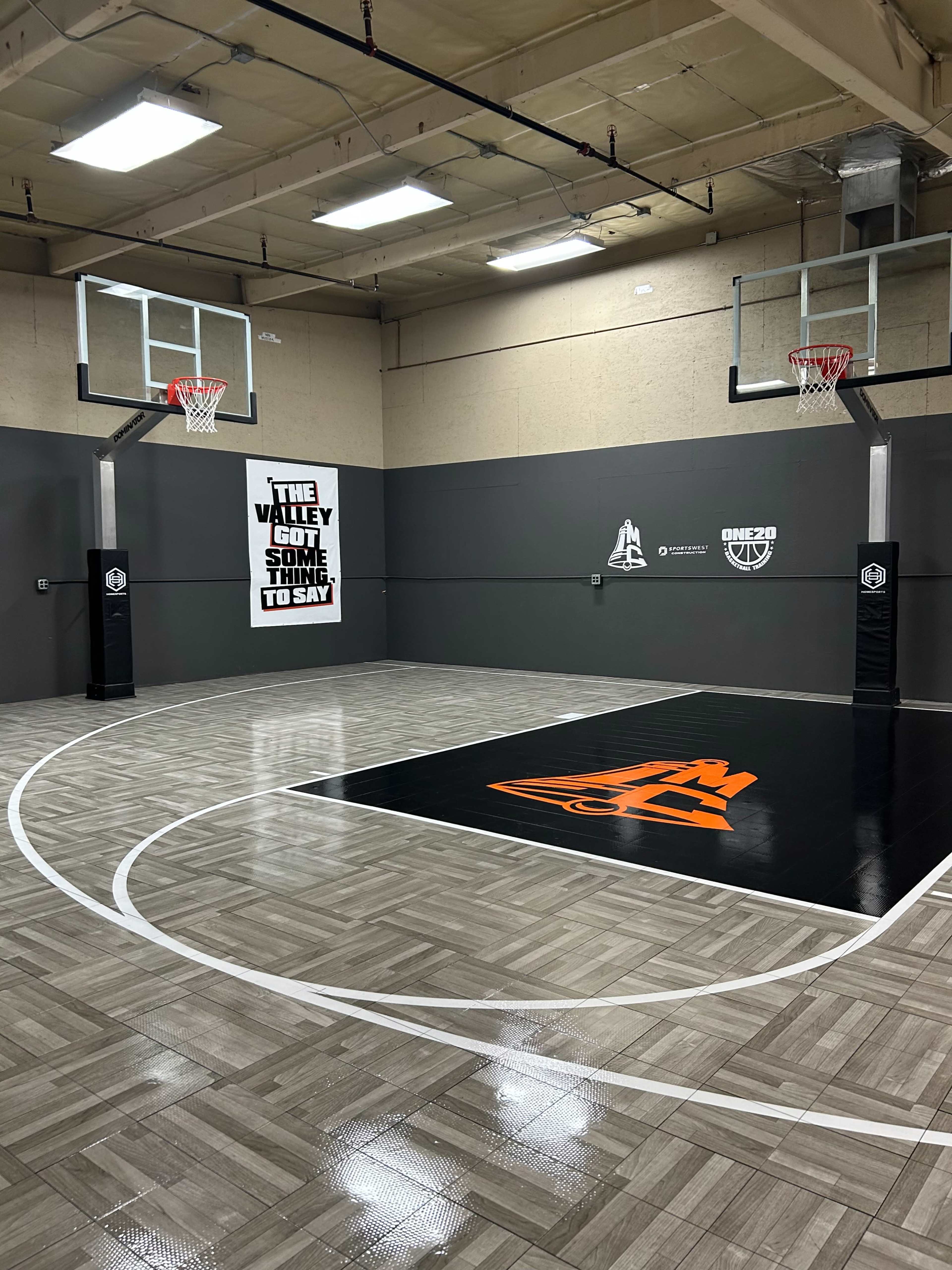 The image shows a basketball court featuring two hoops and a logo at center court, with a gray and black color scheme on the walls.