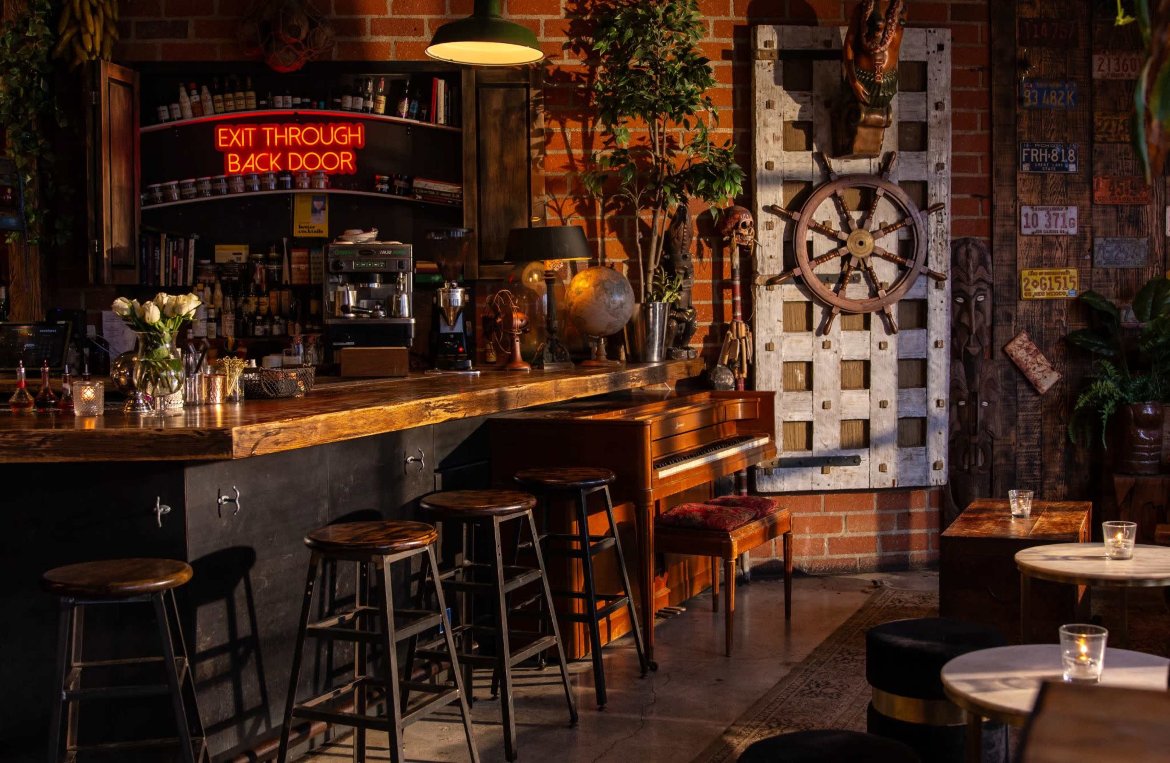 The image shows a cozy bar interior with a wooden counter, a piano, and a mural featuring a nautical wheel against a brick wall, illuminated by soft lighting.