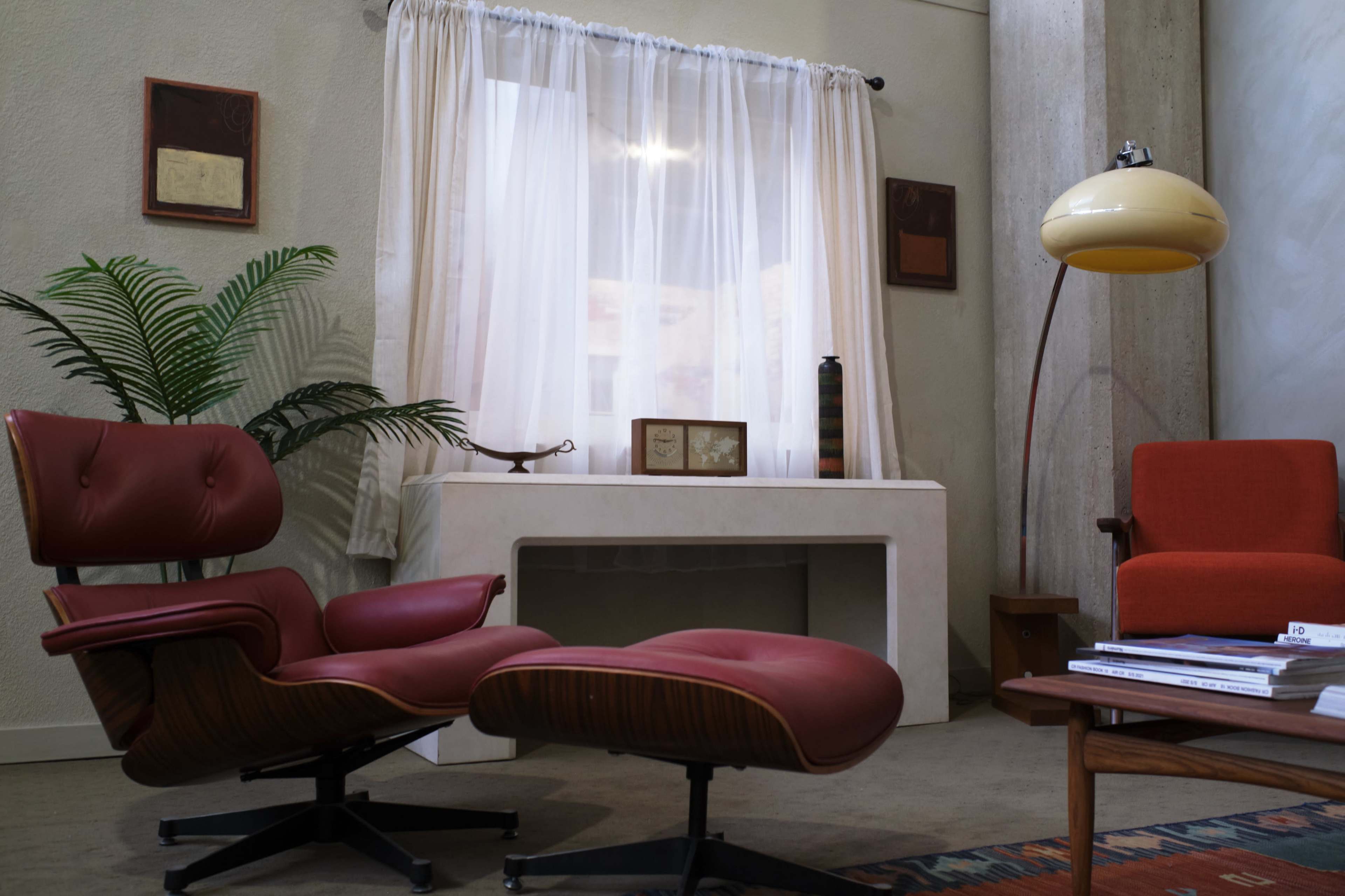 A mid-century modern interior with two red lounge chairs, a white desk, a floor lamp, and a plant near a large window with sheer curtains.