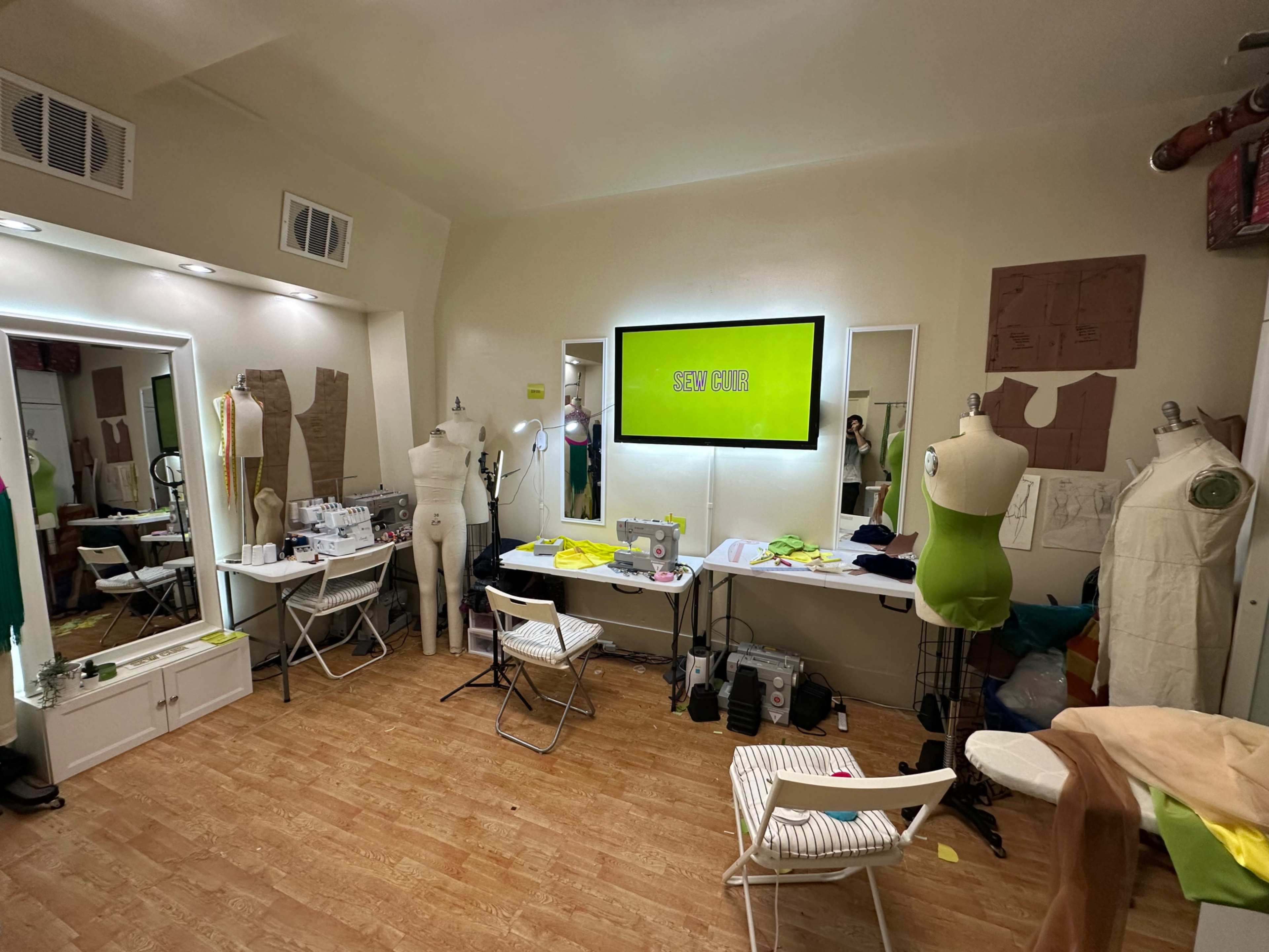 The image shows a sewing studio with mannequins, tables, sewing tools, and bright green signage on the wall.