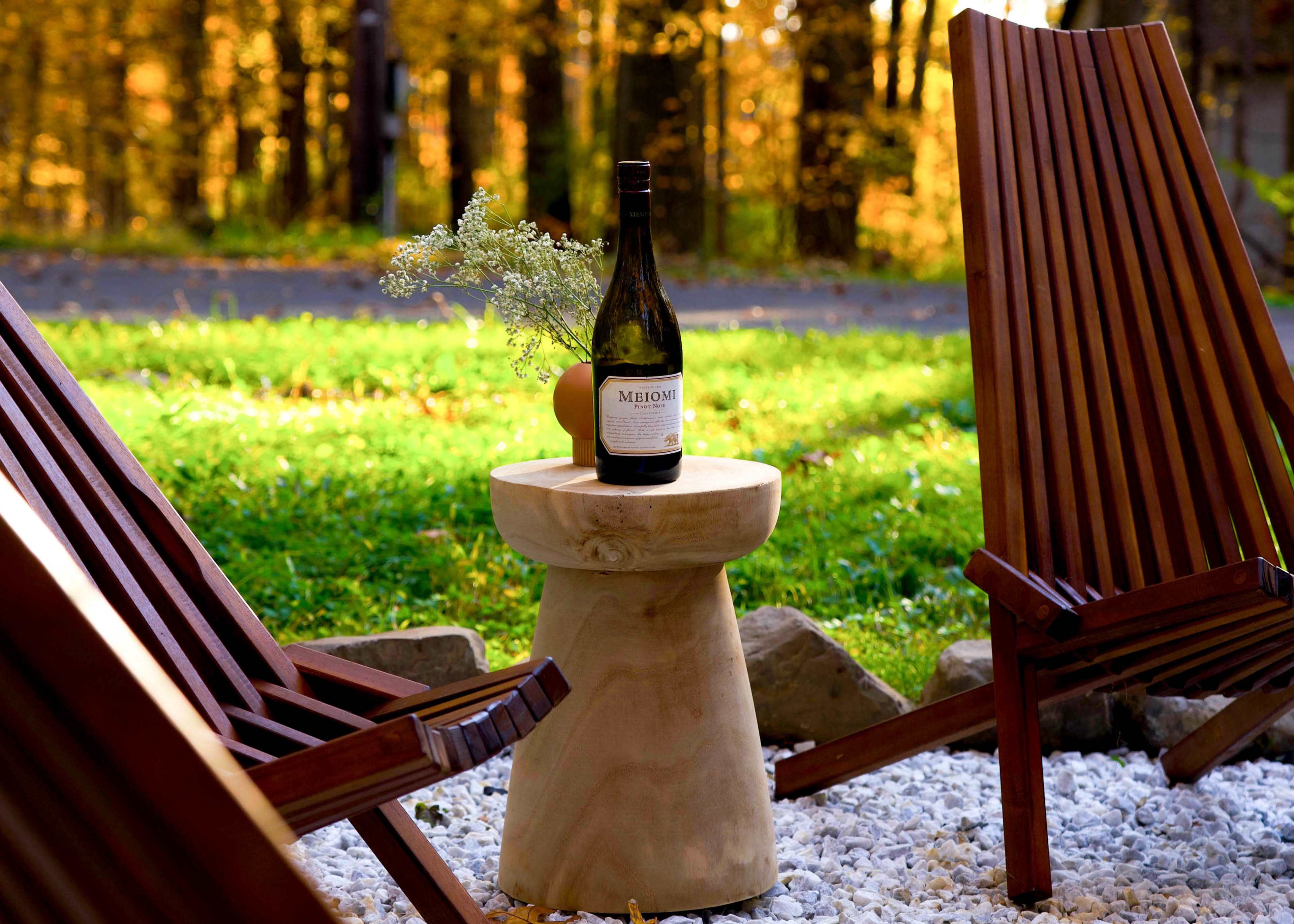 A bottle of wine sits on a wooden stool between two wooden chairs in a green outdoor setting.