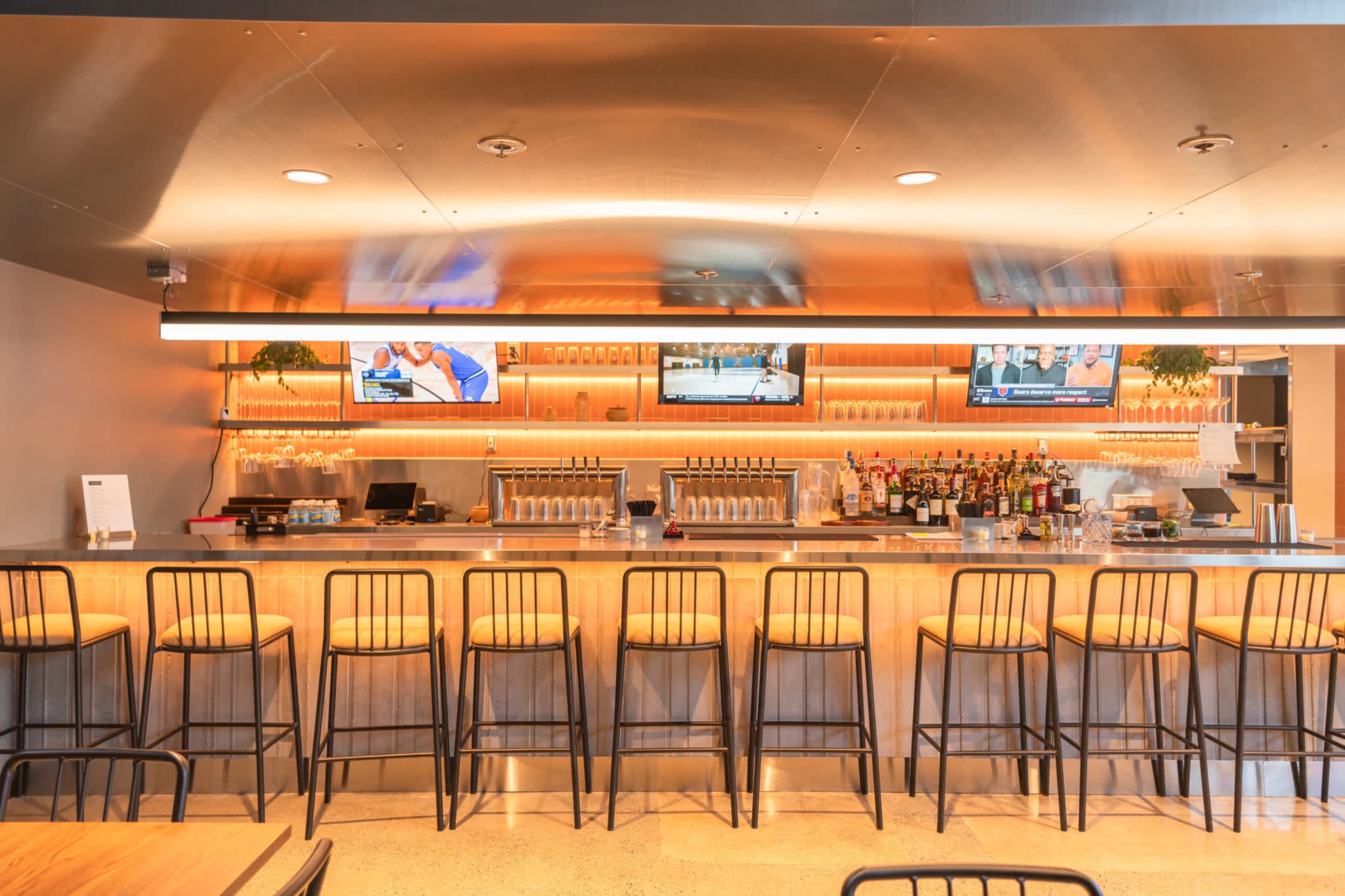 The image shows a modern bar area with a polished countertop, several bar stools, and two television screens displaying sports.