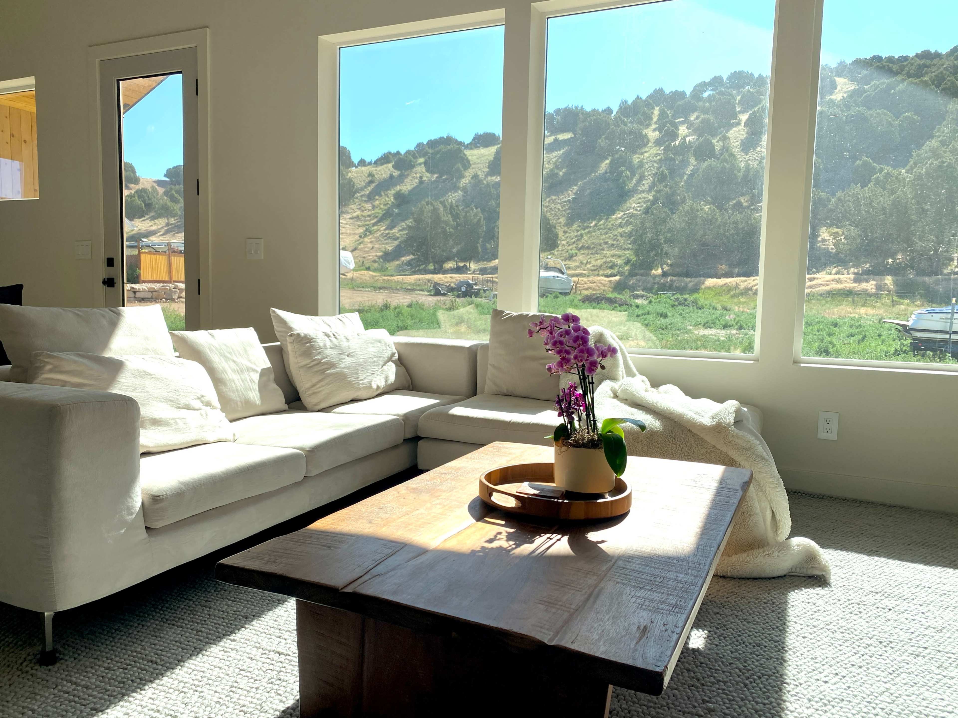 The image shows a modern living room with a white sofa, a wooden coffee table, and large windows that overlook a green landscape with hills.
