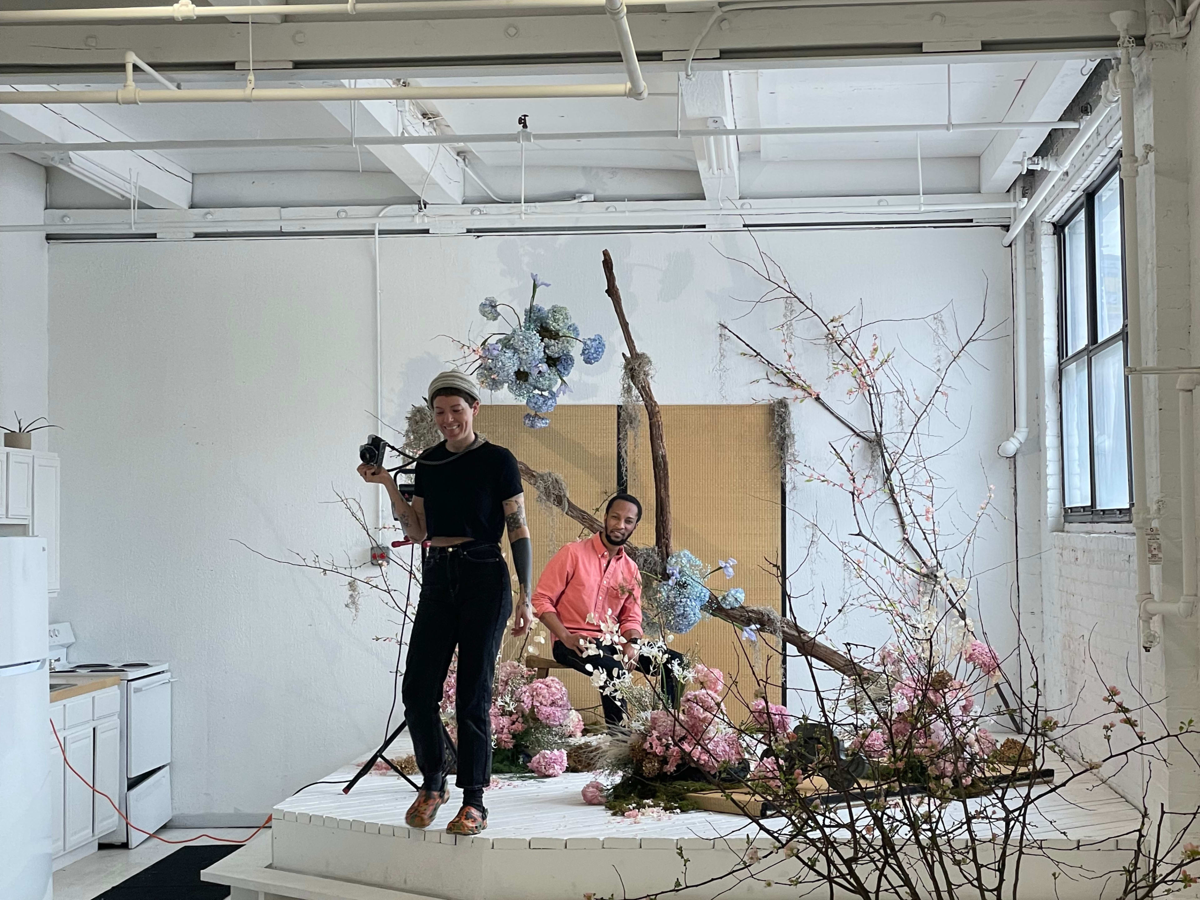 Two individuals in a studio are working on a floral arrangement, with one holding a camera and the other seated among decorative flowers.