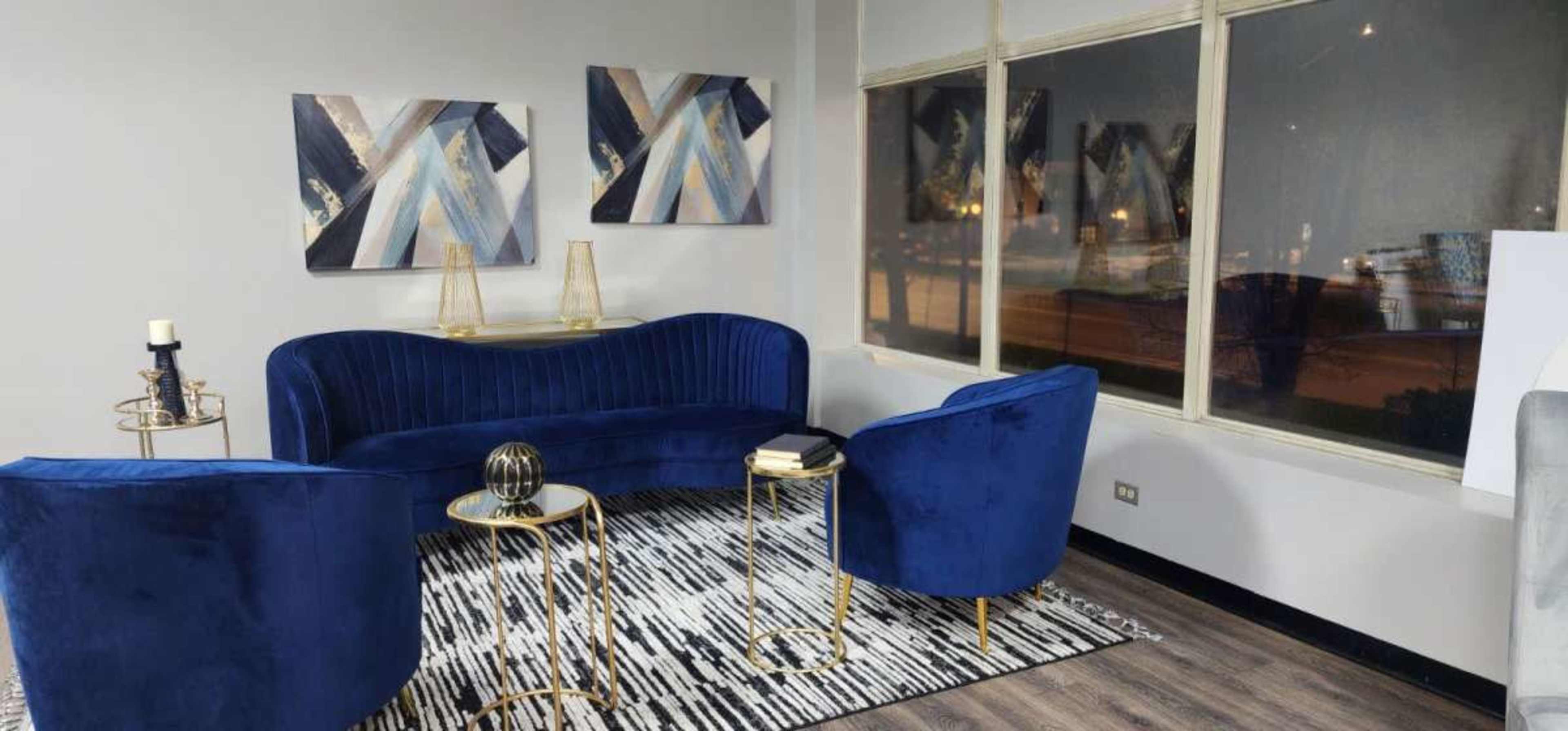 A modern lounge area featuring a blue velvet sofa and two matching chairs, with abstract art on the walls and a patterned rug on the floor.