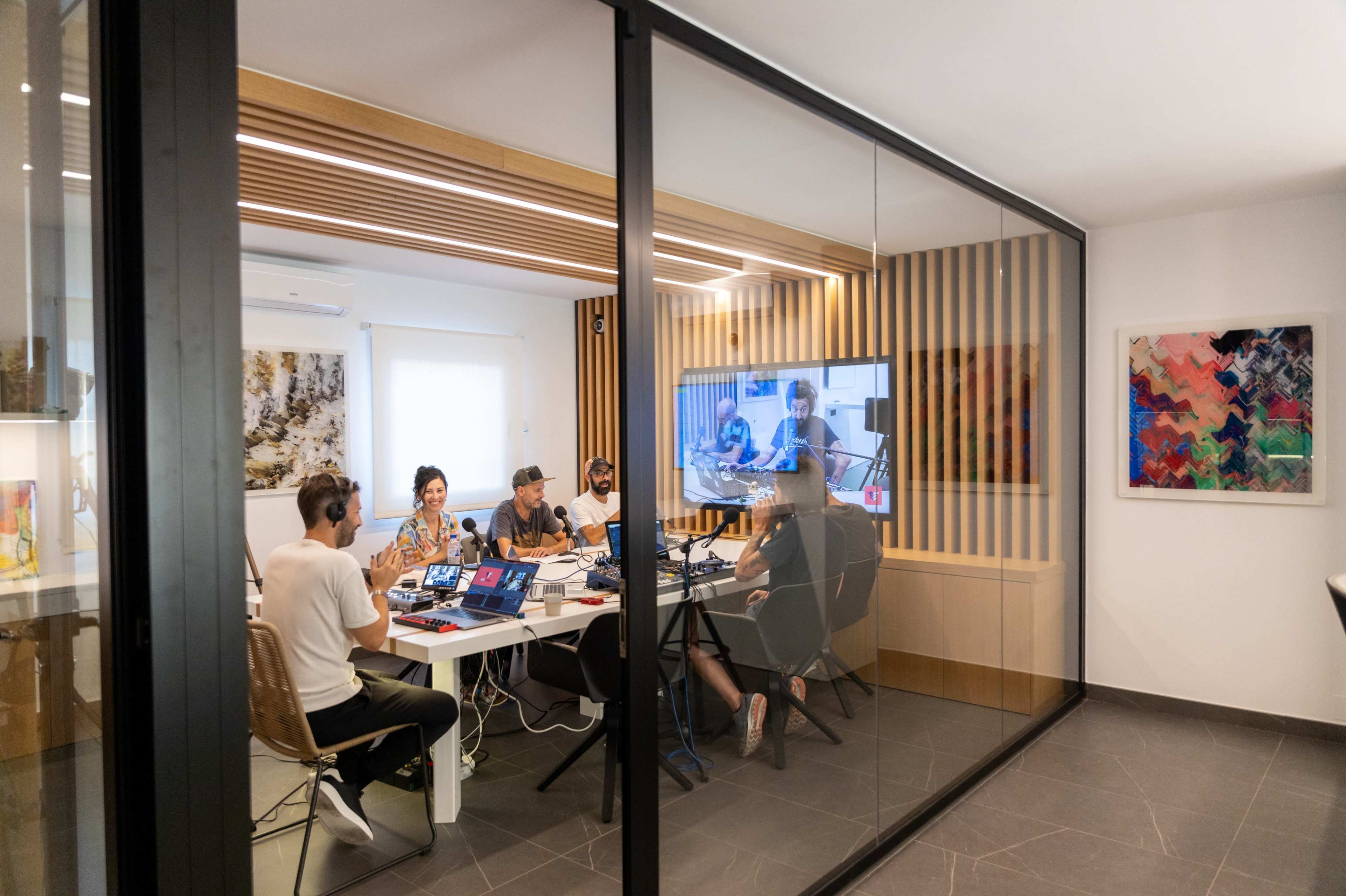 A group of five people is engaged in a video conference in a room separated by glass walls, equipped with computers and audio equipment.