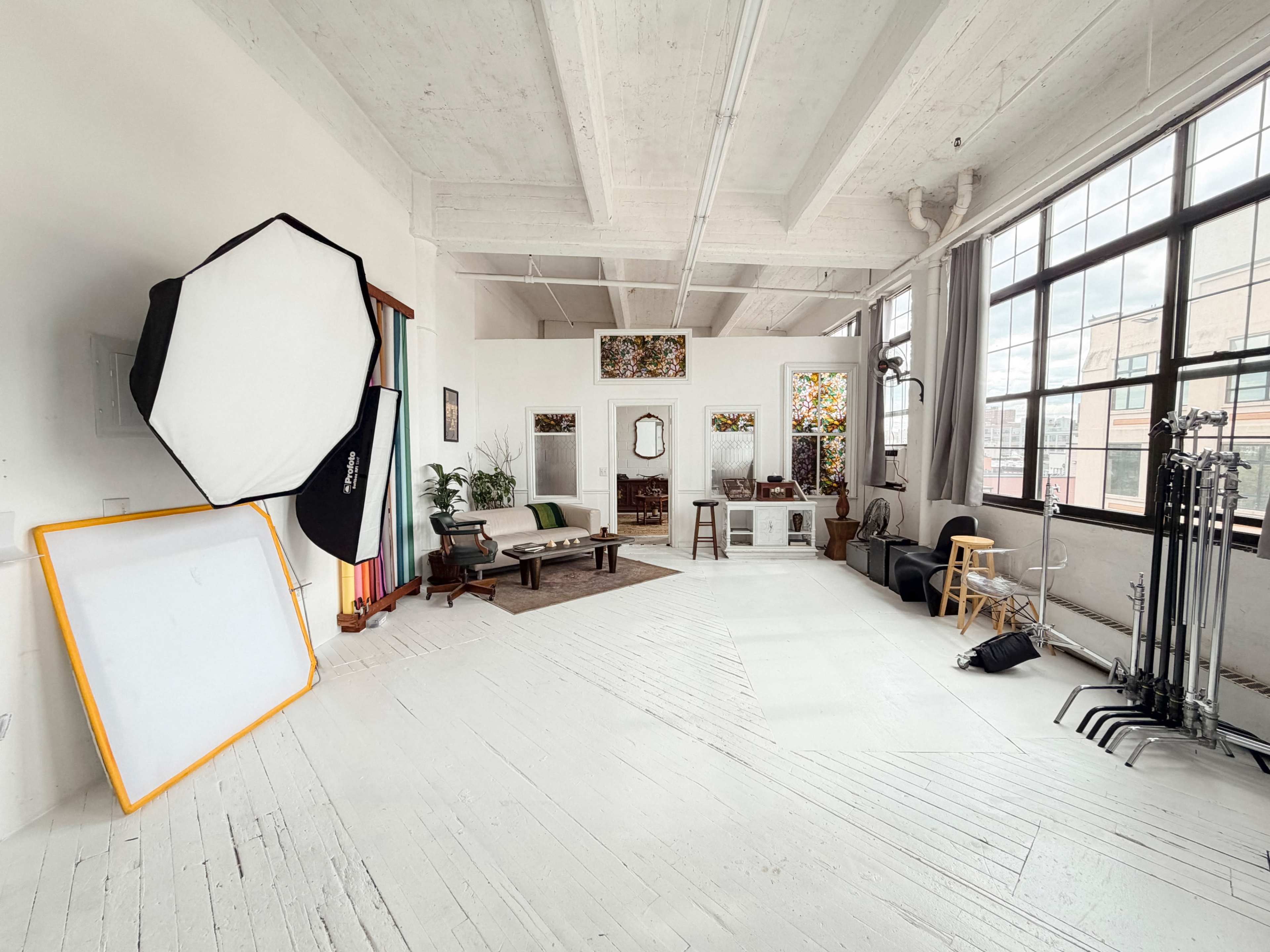 The image shows a bright, spacious studio with large windows, featuring various furniture pieces, a reflector, a softbox light, and a backdrop setup.