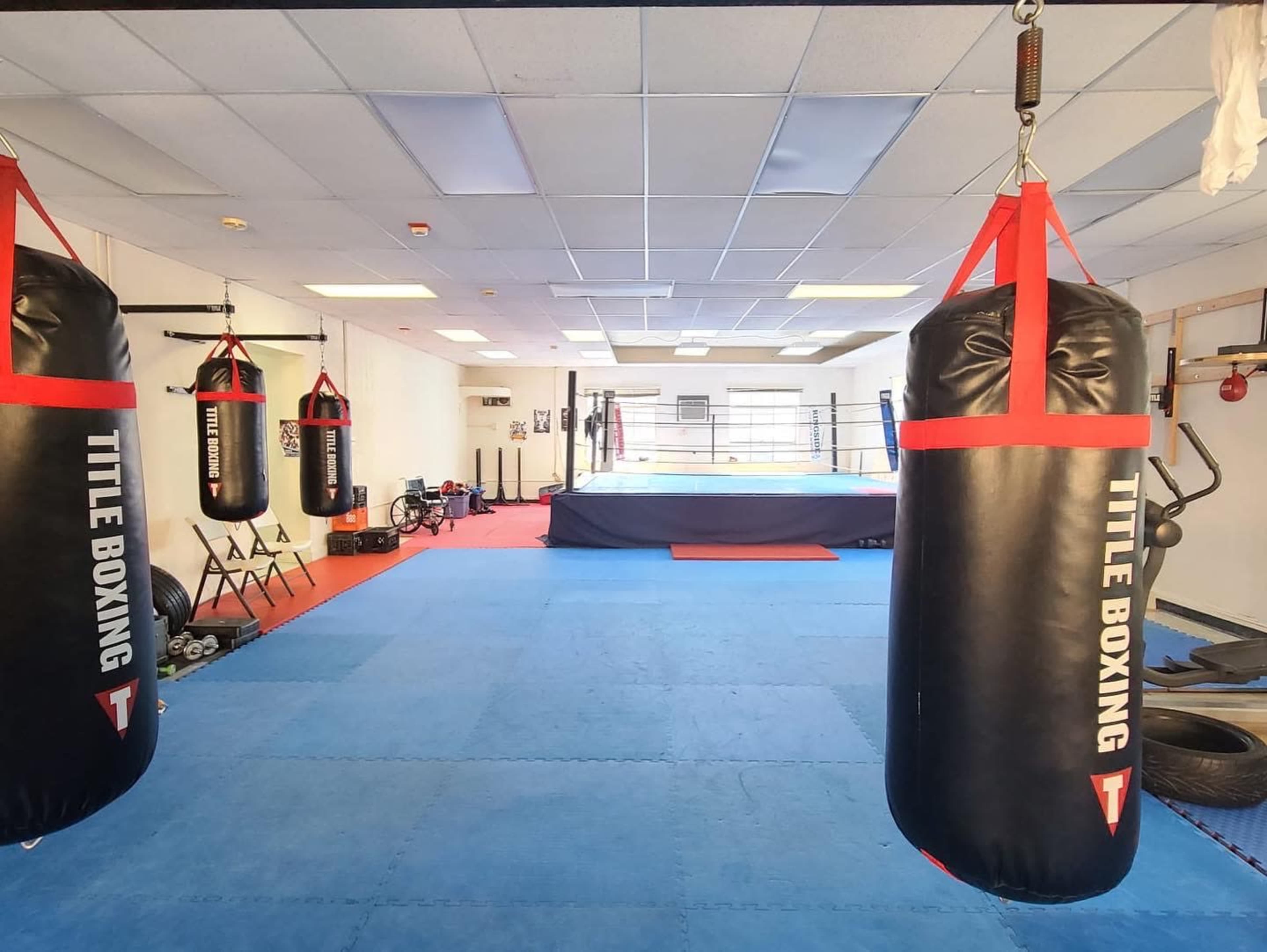 A boxing gym with several heavy bags hanging, blue mats on the floor, and a boxing ring in the background.