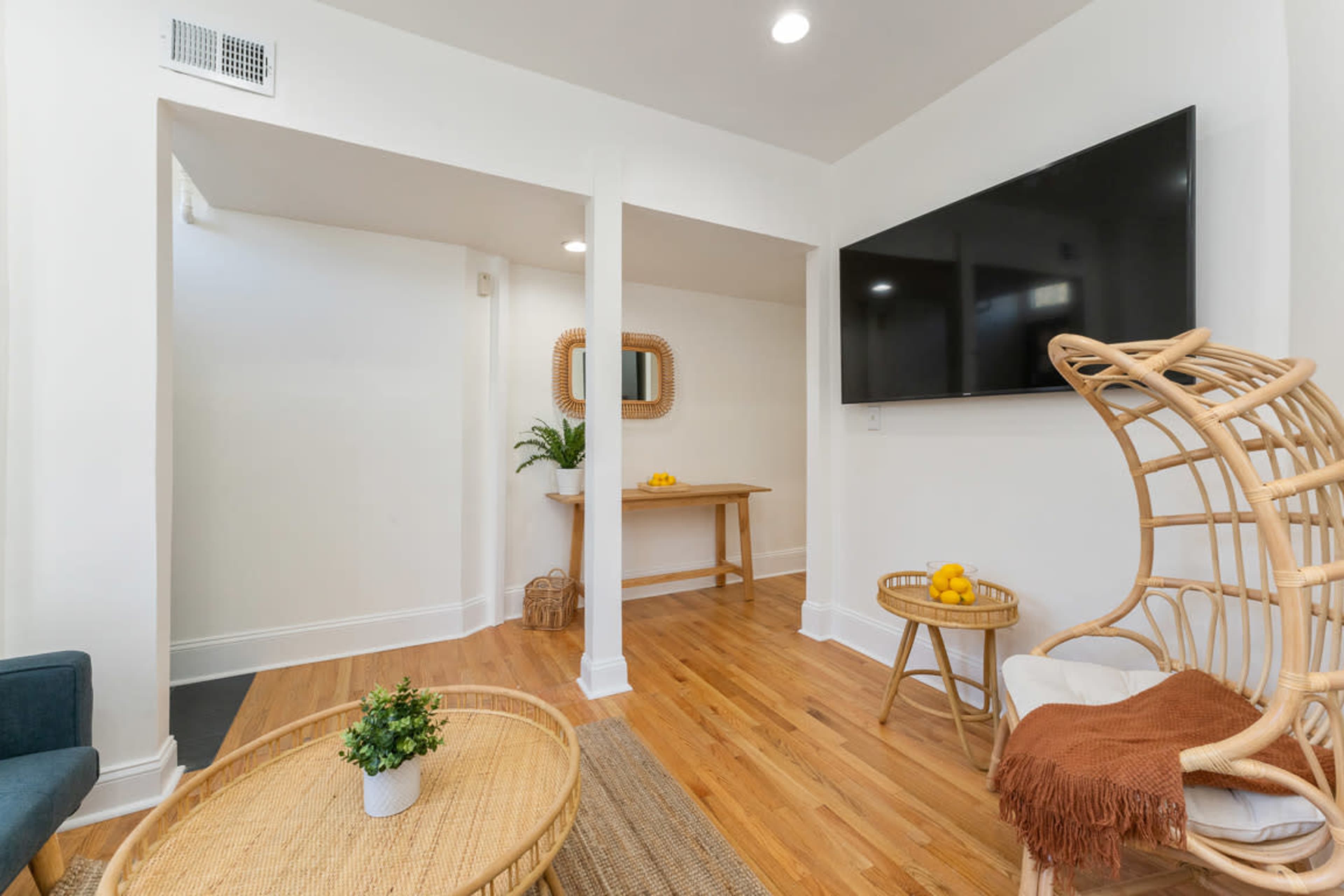 The room features a light-colored, minimalist interior with a large TV mounted on the wall, a wooden table, a rattan chair, and decorative plants.