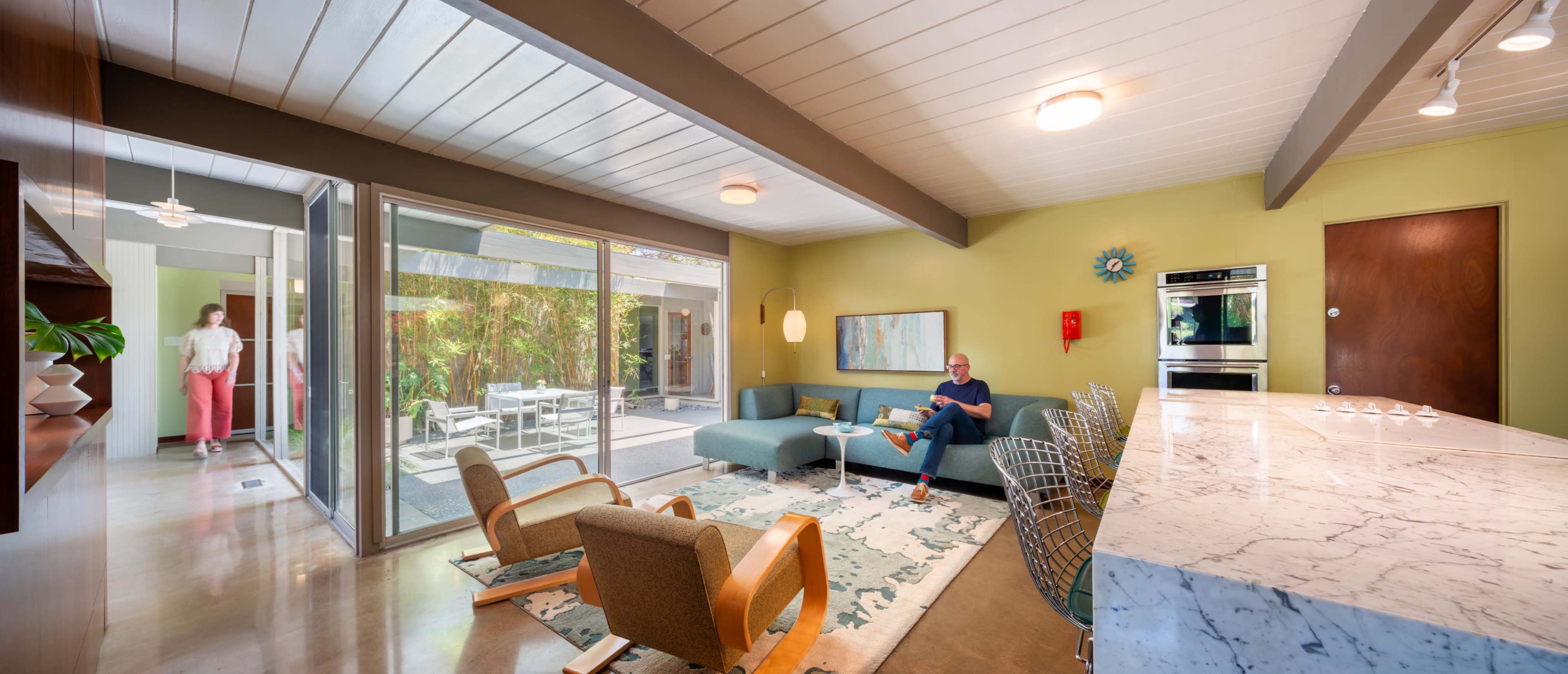 The image shows a modern living room with large windows, a view of an outdoor space, and two people seated in the room.