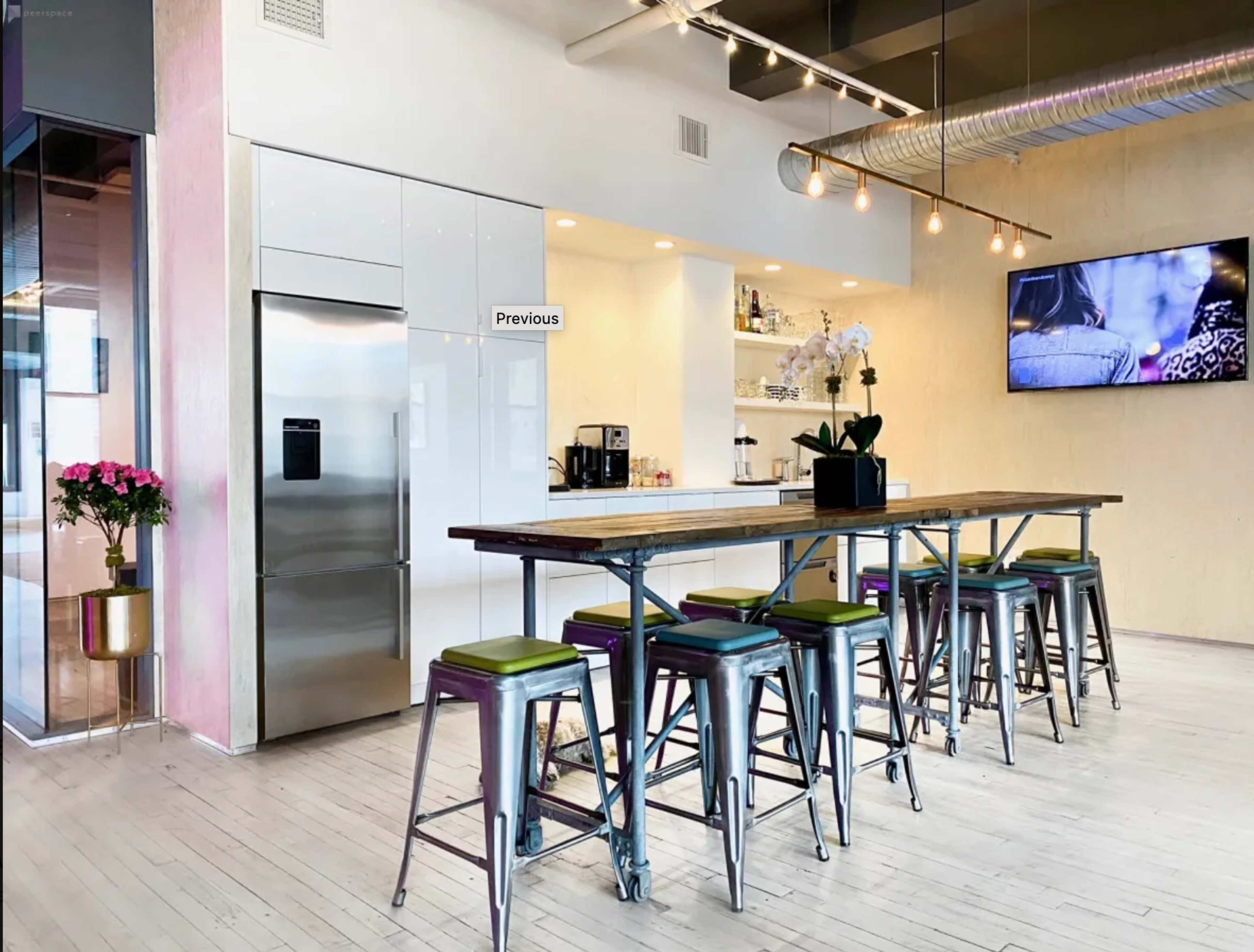 A modern kitchen area with a long wooden table surrounded by metal stools, a refrigerator, and a beverage station under soft lighting.