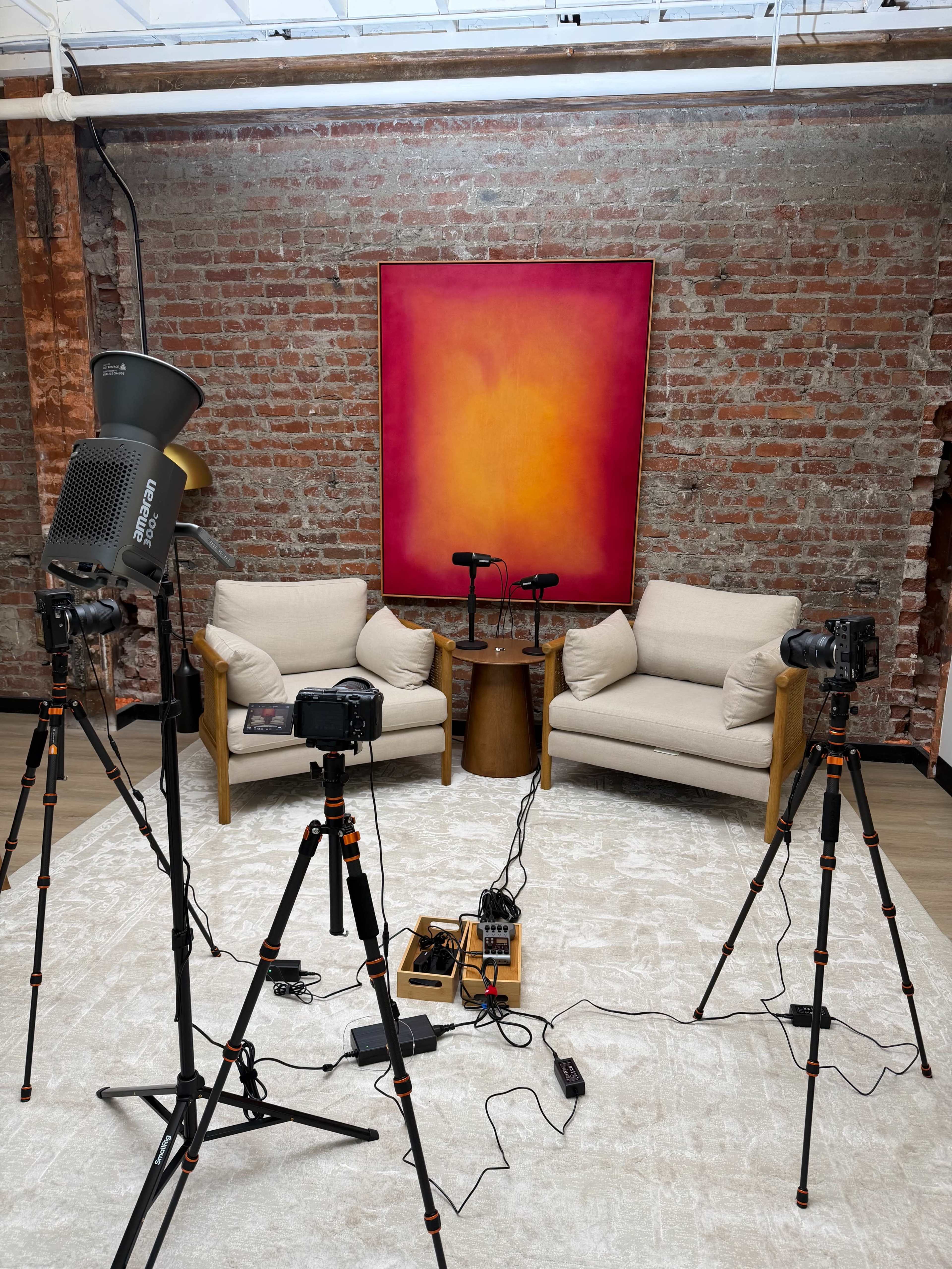 A spacious room features two light-colored chairs facing each other, a small wooden table between them, and several cameras and microphones set up on tripods against a backdrop of exposed brick and a colorful abstract painting.