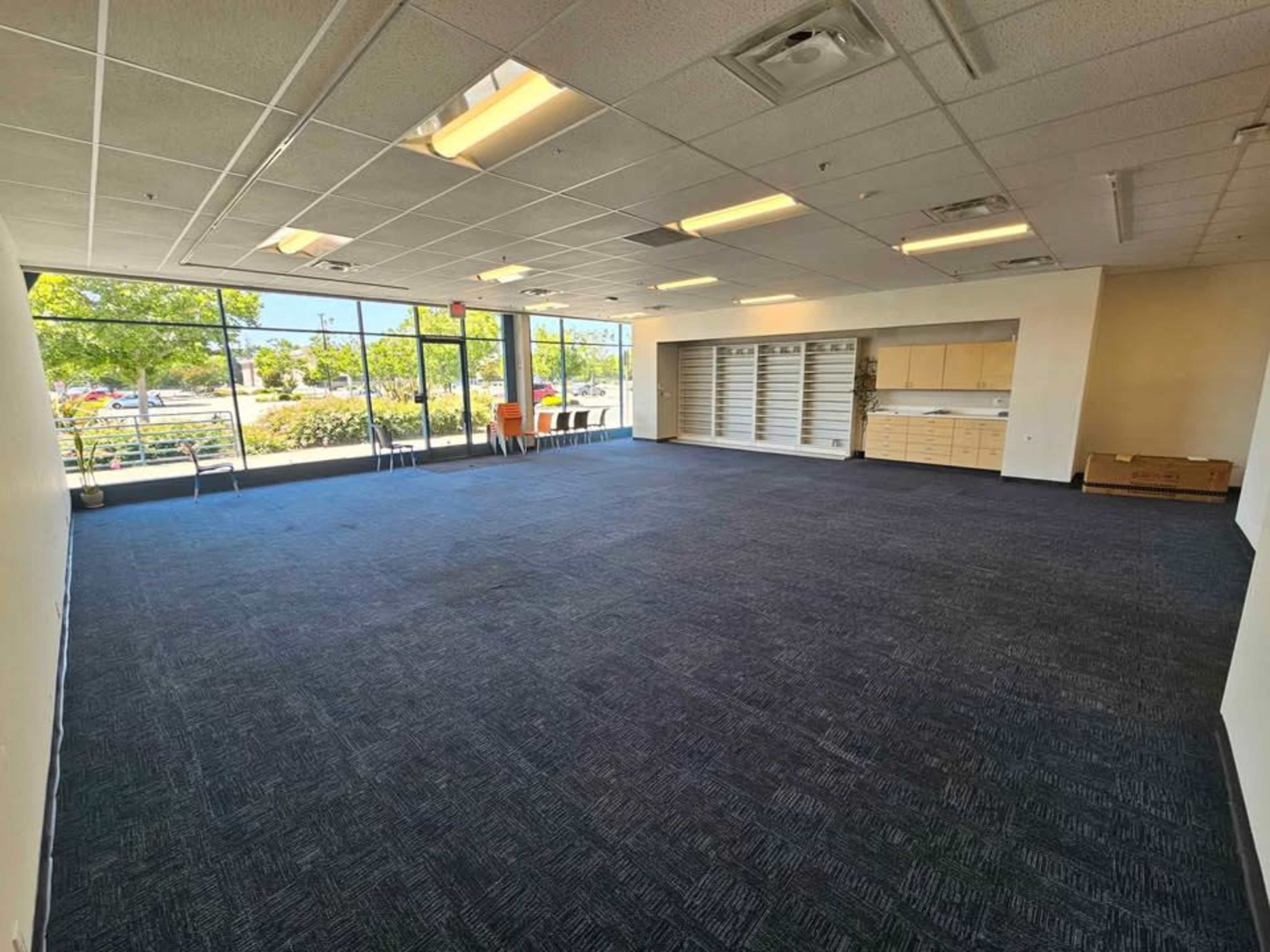 The image shows a spacious, empty classroom with blue carpet, large windows letting in natural light, and built-in storage cabinets along one wall.