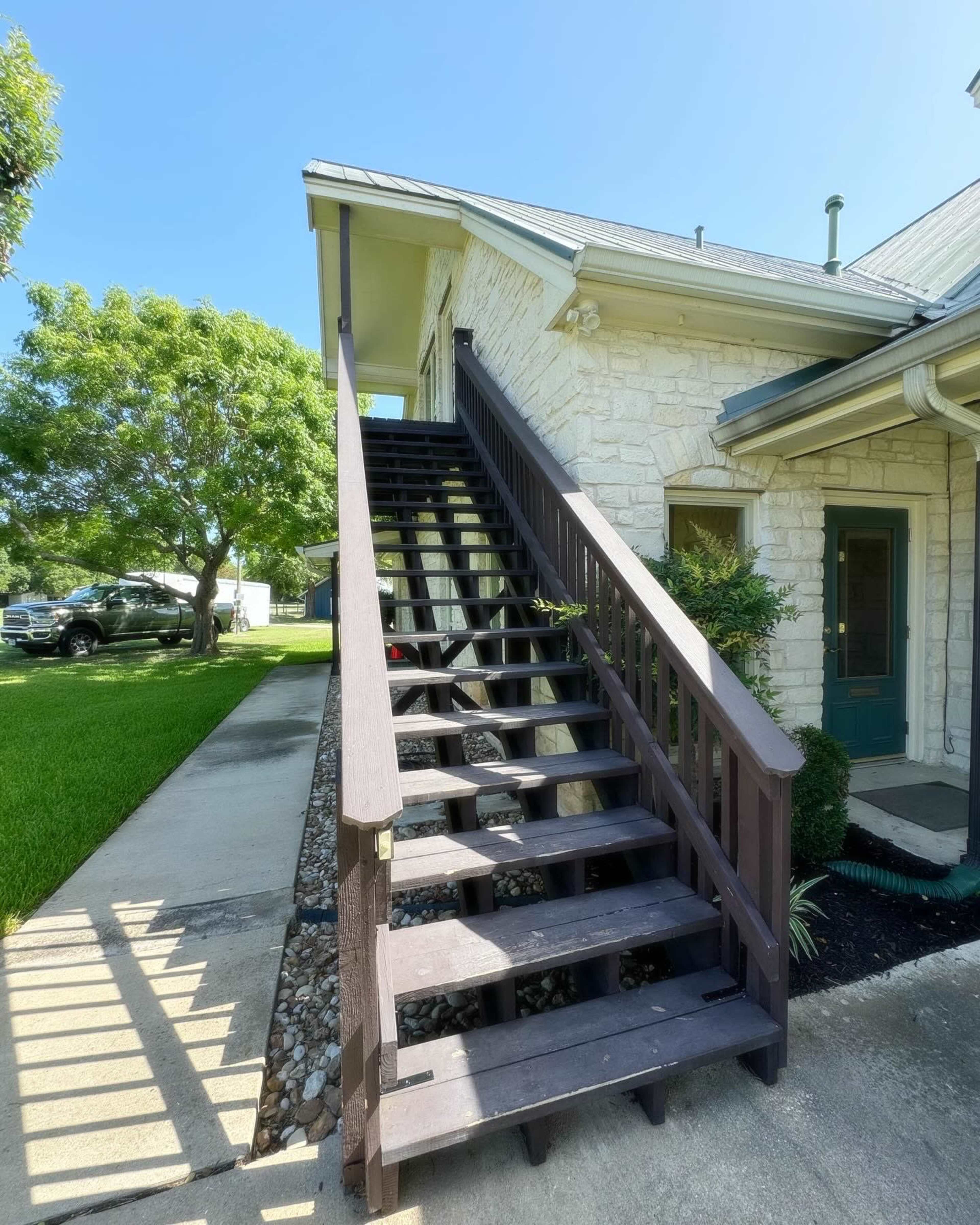 A set of wooden stairs leading to a second-story entrance of a house, with a grassy area and parked vehicle nearby.