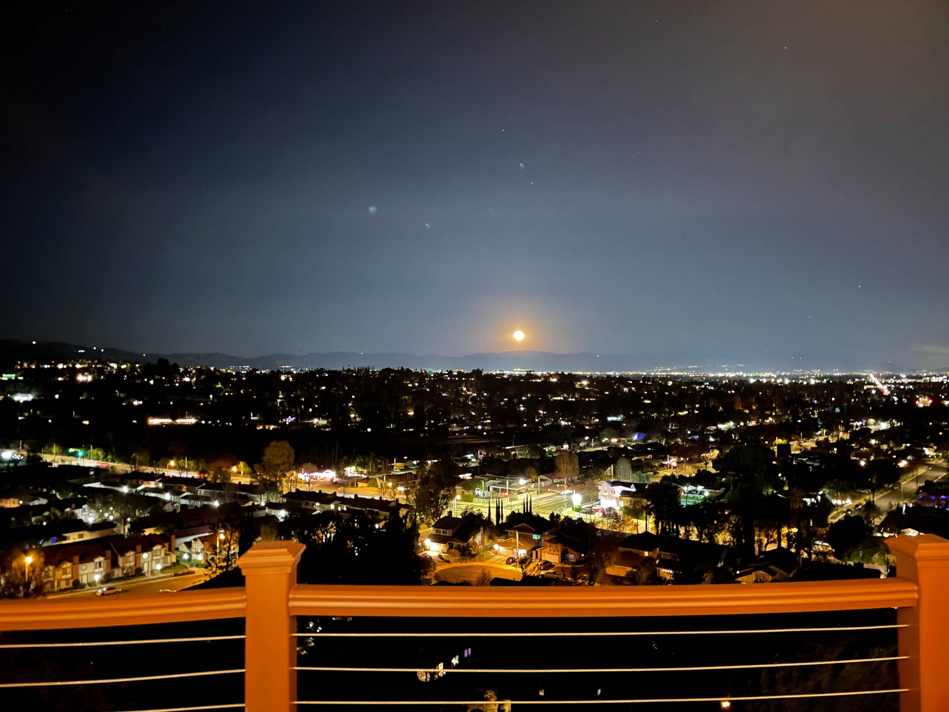 A full moon rises over a cityscape illuminated by numerous lights at night.