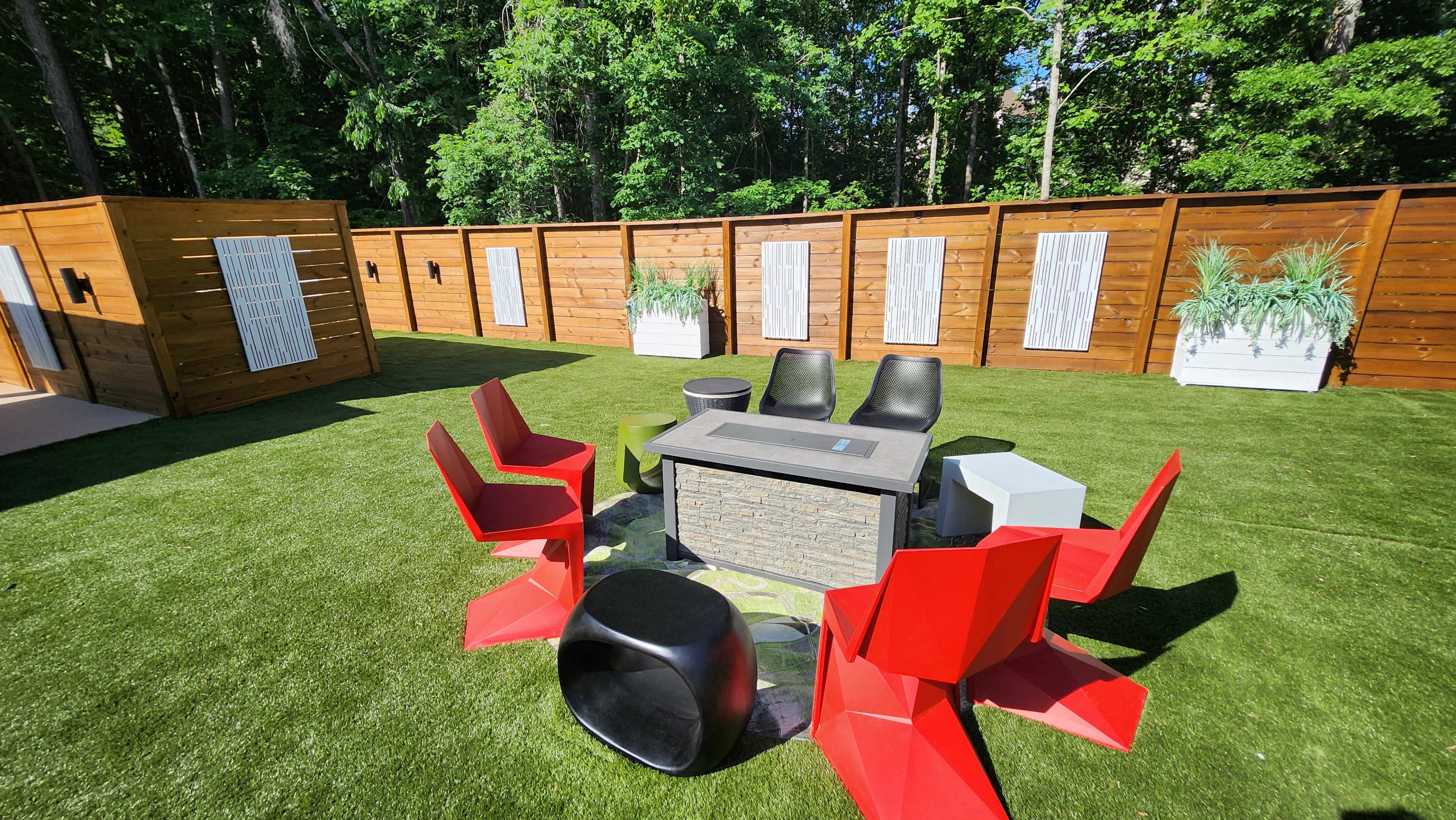 A modern outdoor seating area features a stone table, black and red chairs, and artificial grass, surrounded by wooden partitions with white accents.