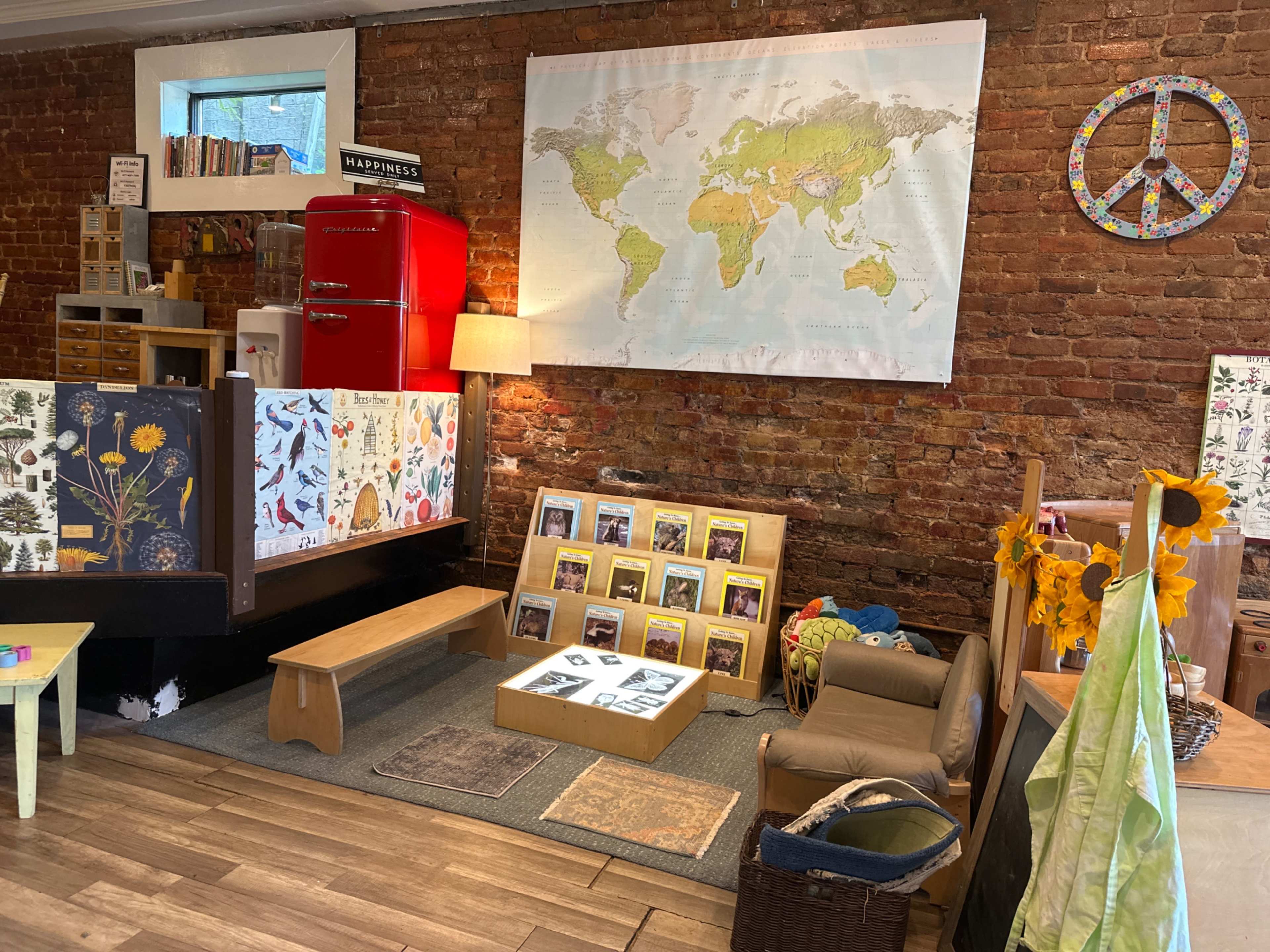 The image shows a cozy interior with brick walls, featuring a world map, a vintage red refrigerator, shelves filled with books, a display of framed artworks, and a seating area with plants and decor.