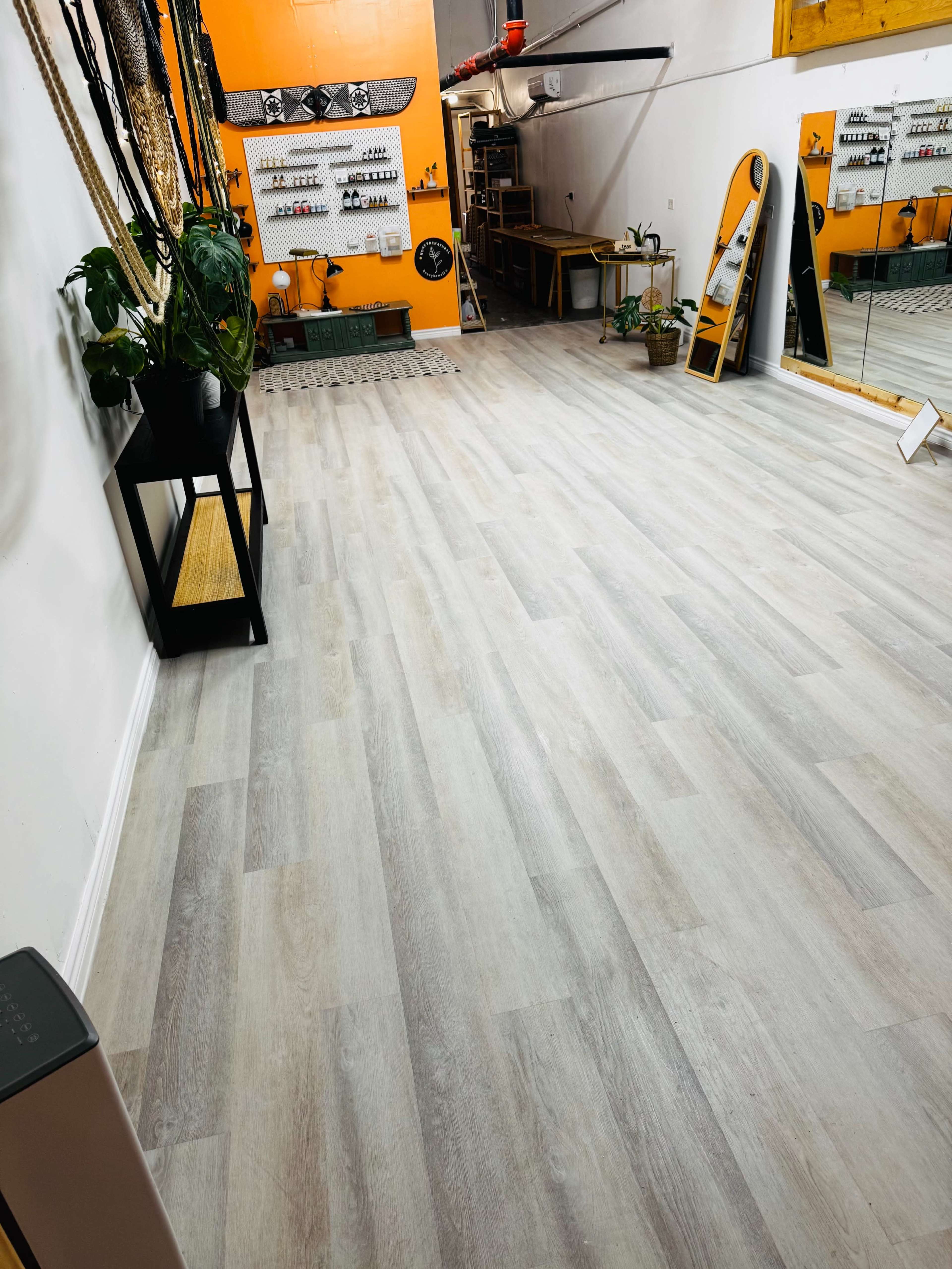 The image shows a spacious interior with light gray wooden flooring, a large mirror, and various items mounted on the wall.