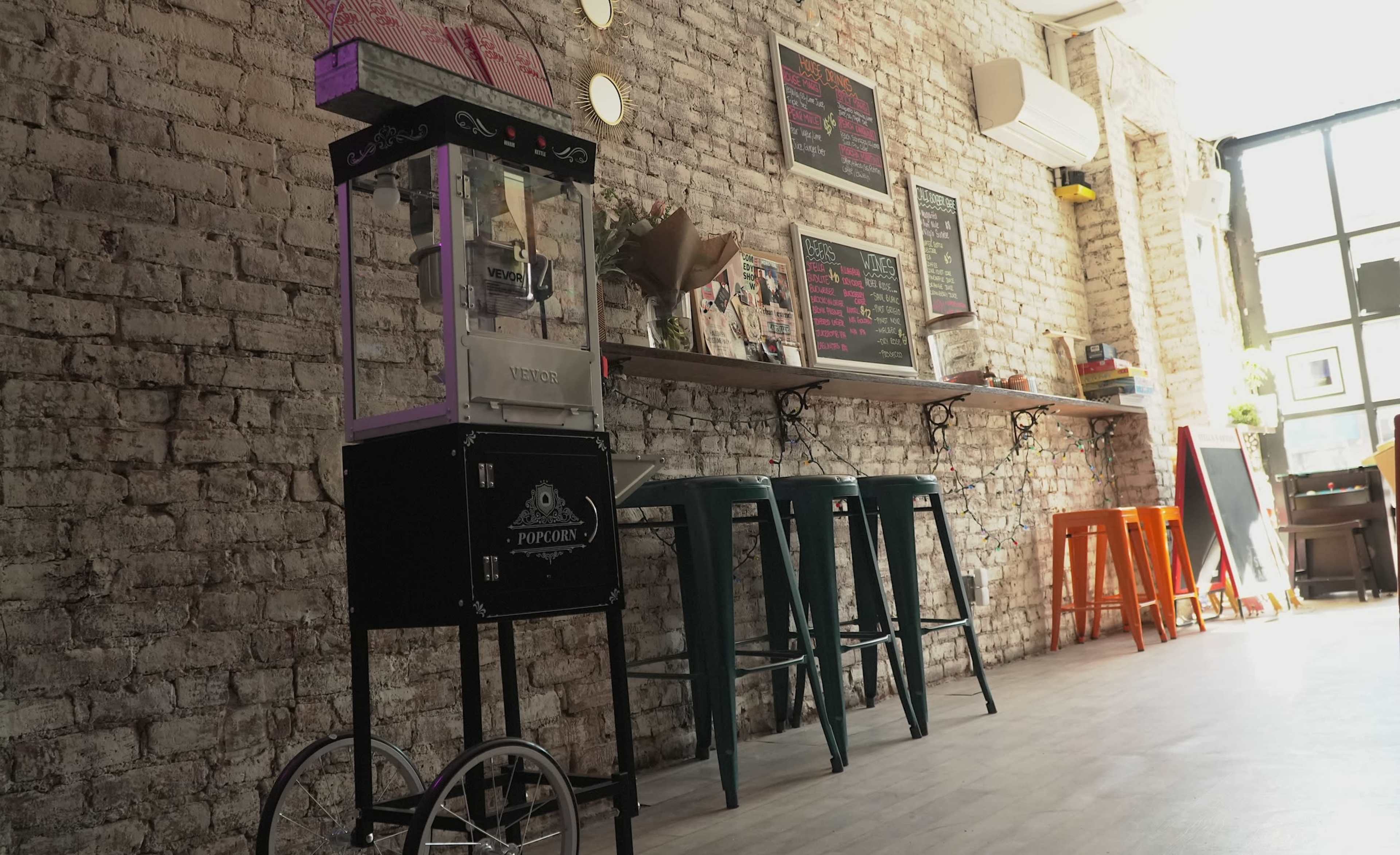 A popcorn machine stands next to colorful stools in a coffee shop with exposed brick walls and a menu displayed on chalkboards.