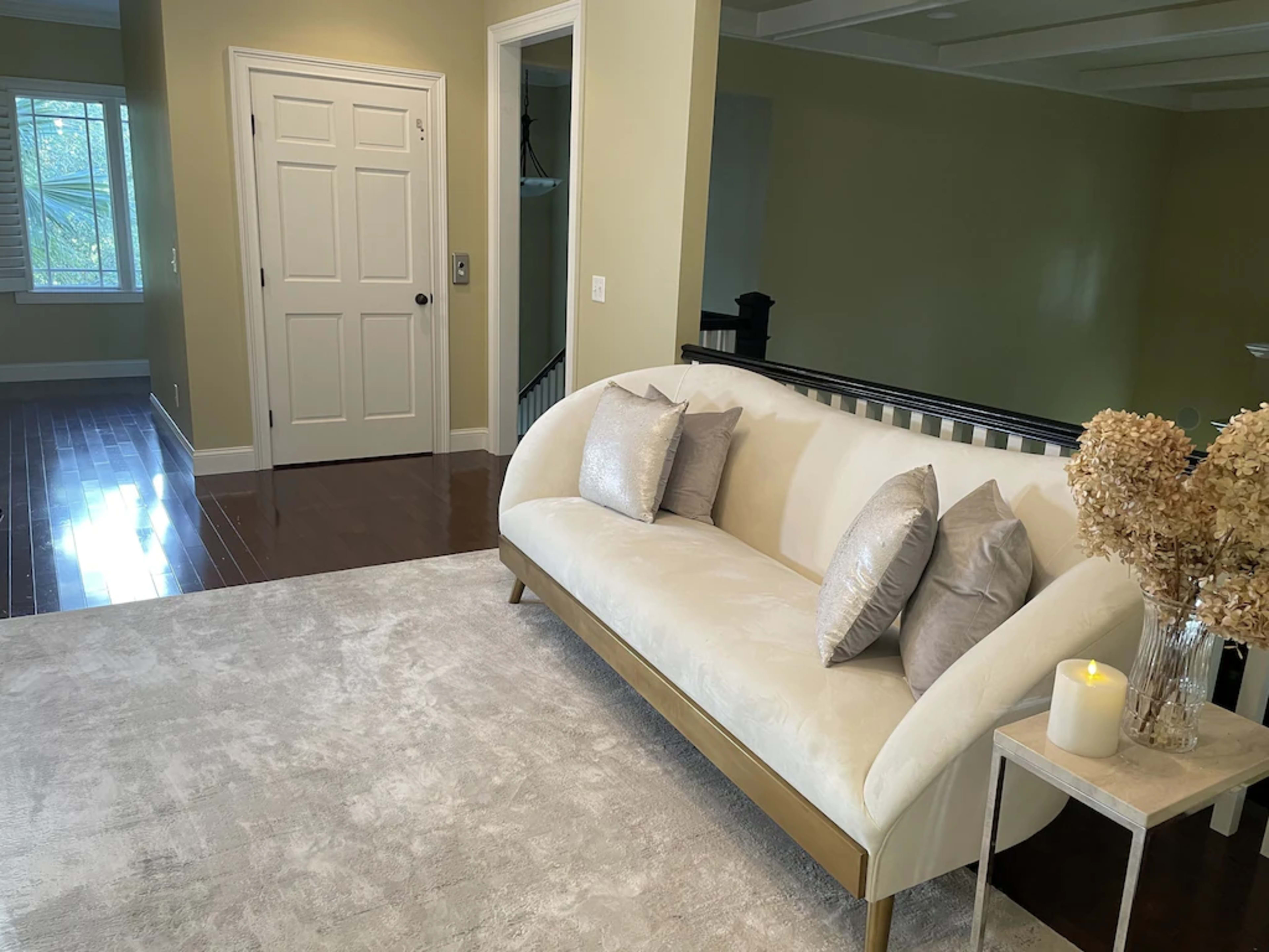 A light-colored sofa with decorative pillows placed on a plush rug in a well-lit room, with a doorway and a staircase visible in the background.