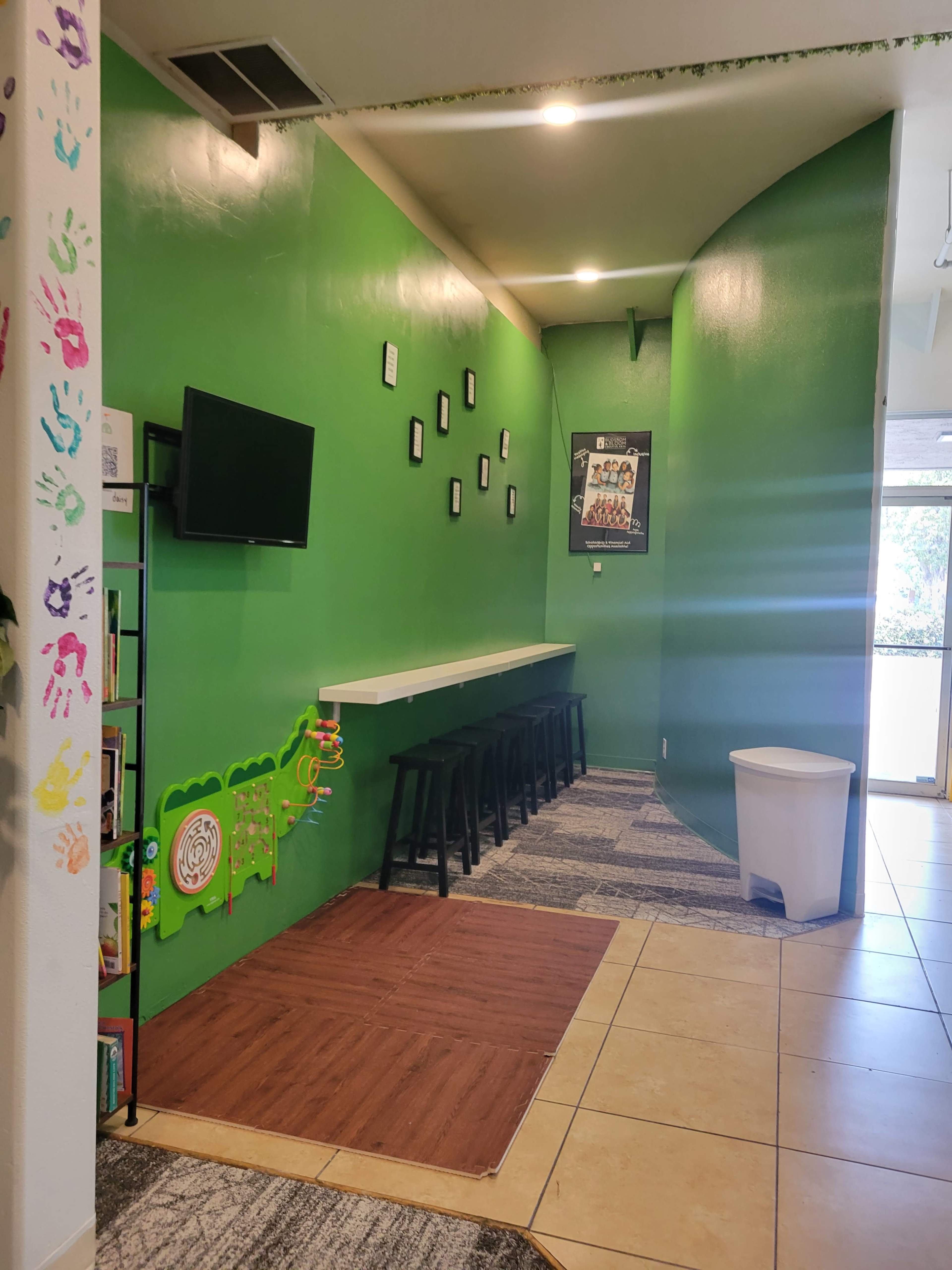 The image shows a small, brightly colored green room with a wall-mounted TV, a long table with stools, and a doorway leading to the outside.