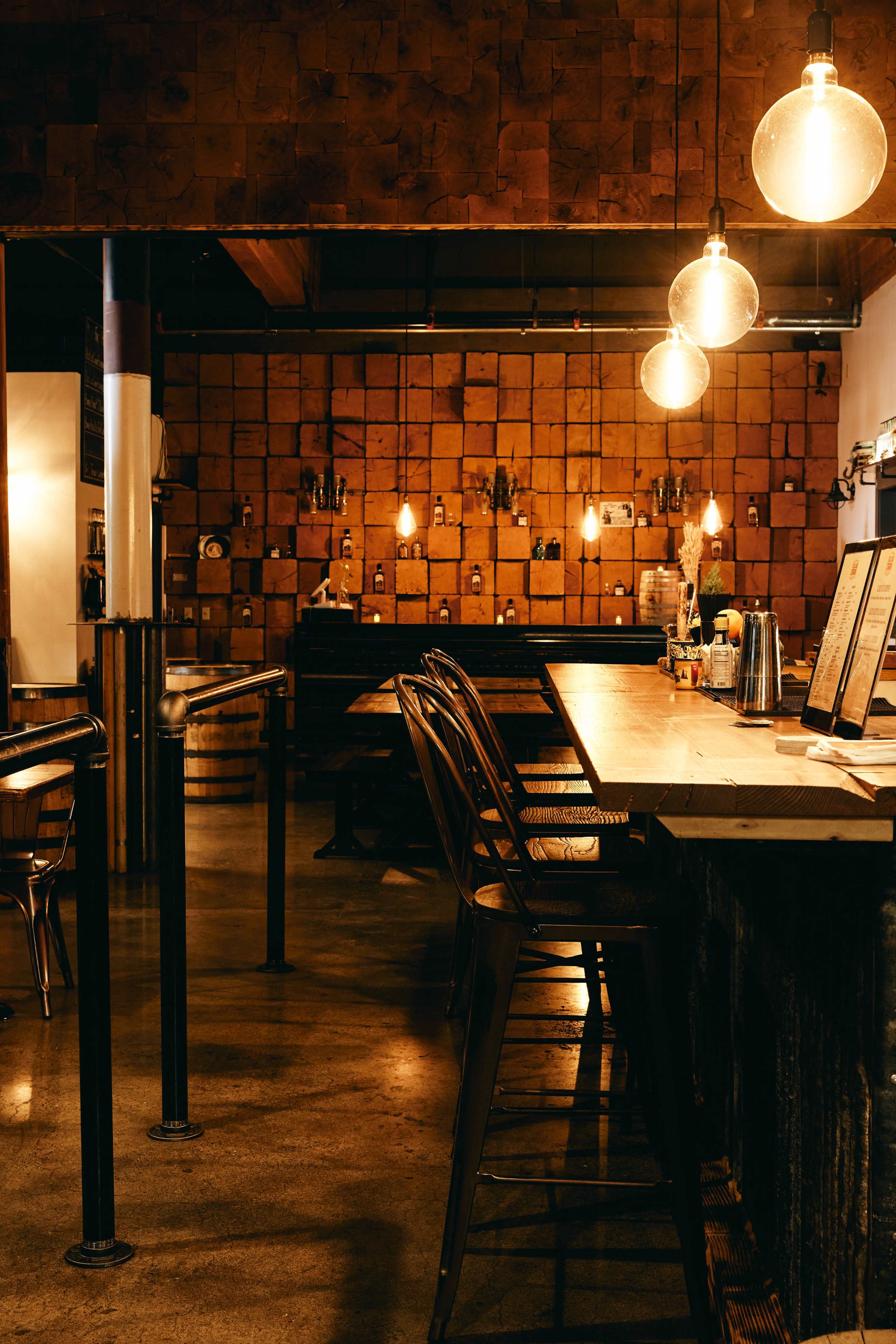 The image shows a dimly lit bar area with wooden walls, metal stools, and hanging light bulbs.