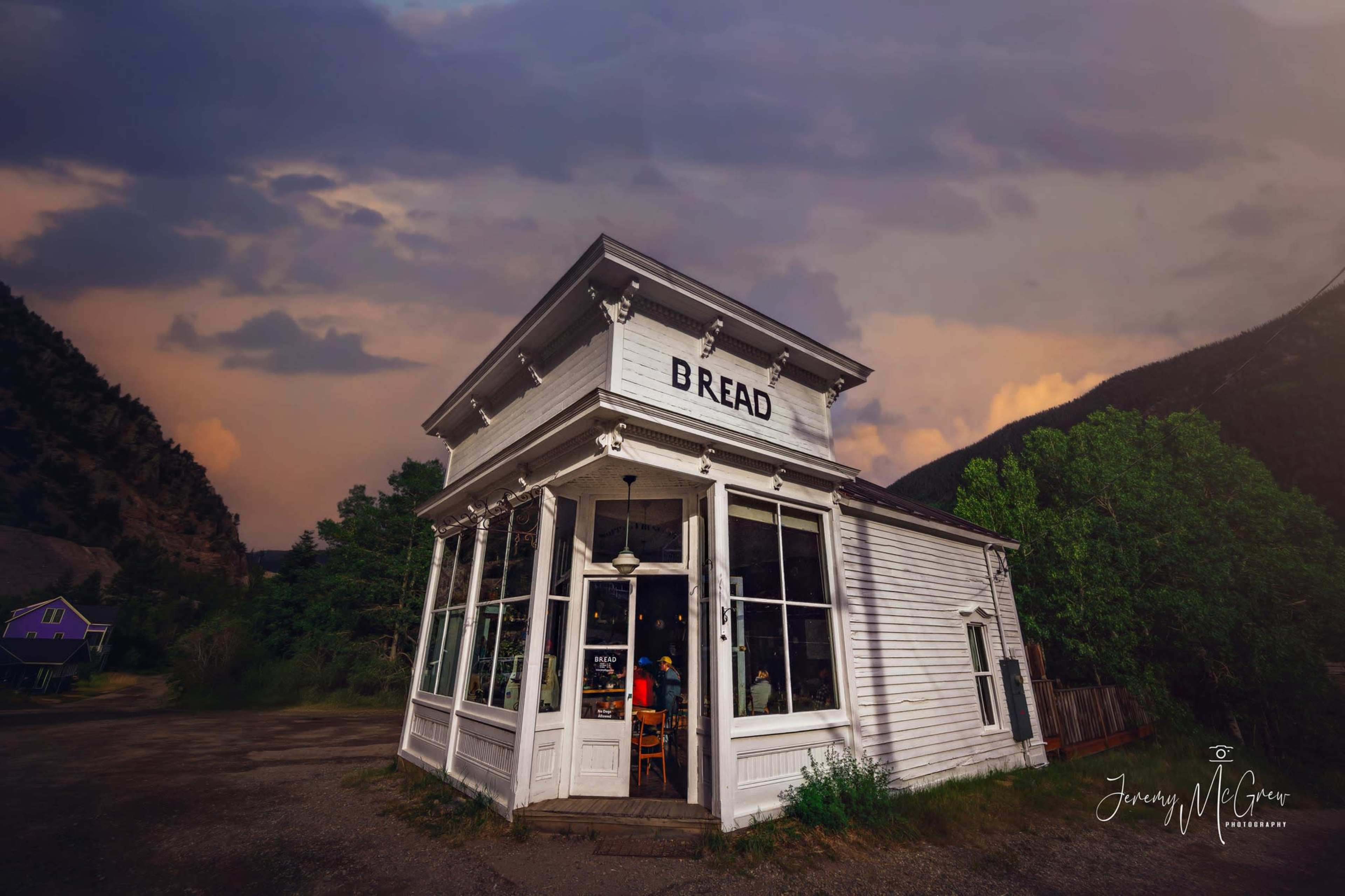 The image shows a white, vintage-style building labeled "BREAD," situated in a mountainous area under a cloudy sky.