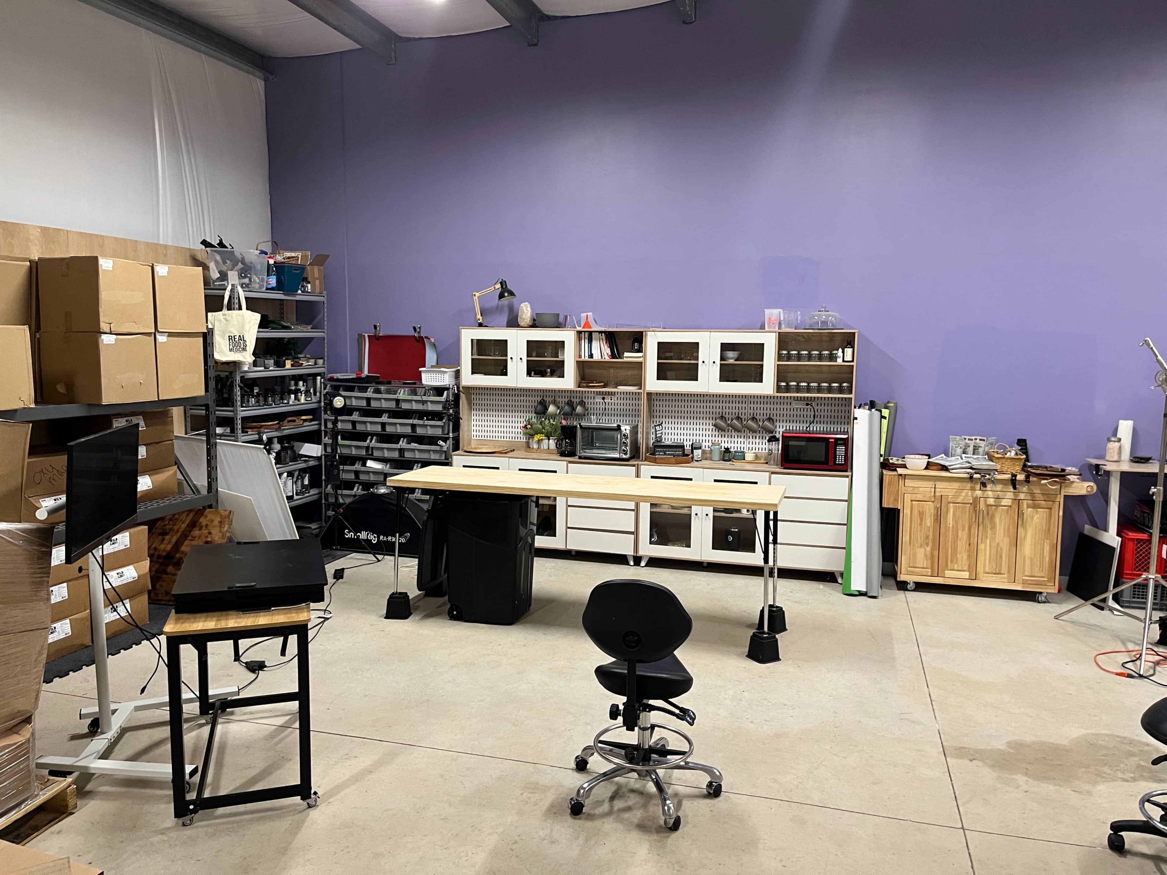 A workspace featuring a large, central table, a black rolling chair, shelving units filled with supplies, and stacks of boxes against the wall.