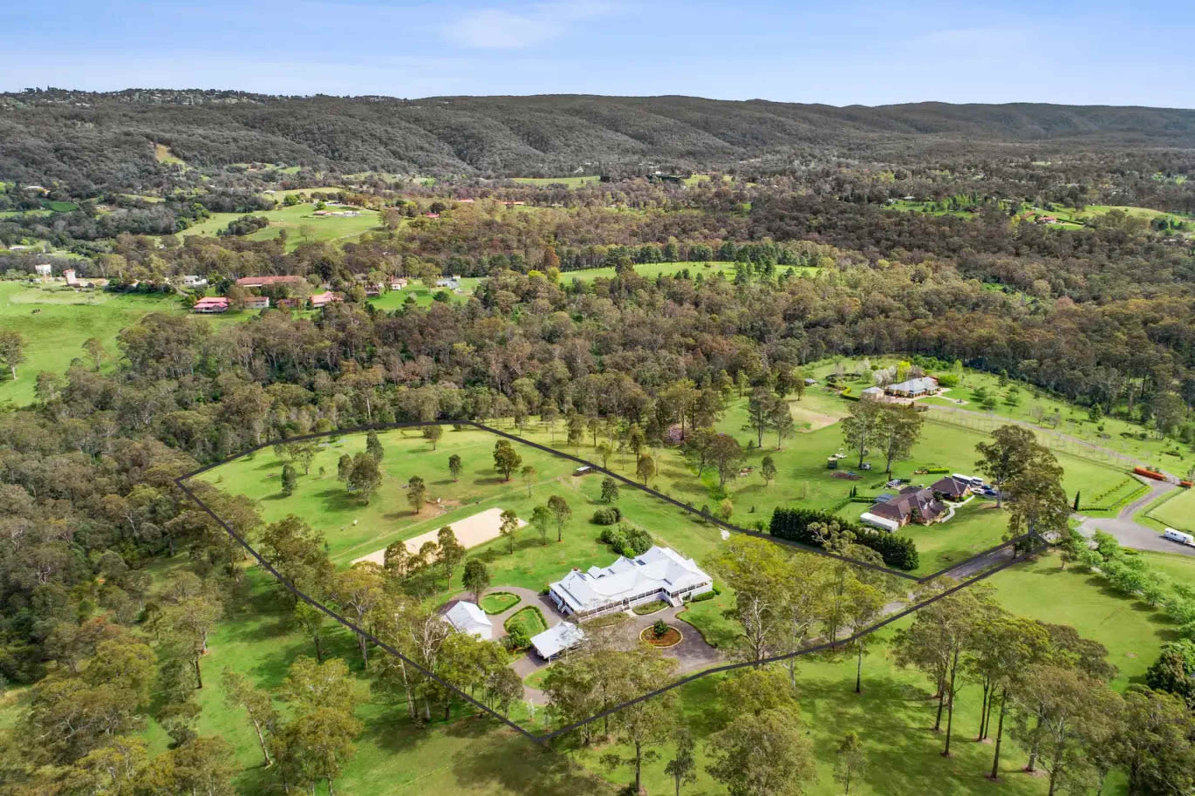 The image shows a sprawling estate surrounded by wooded areas and rolling hills, with a house and several outbuildings set on a large, green property.