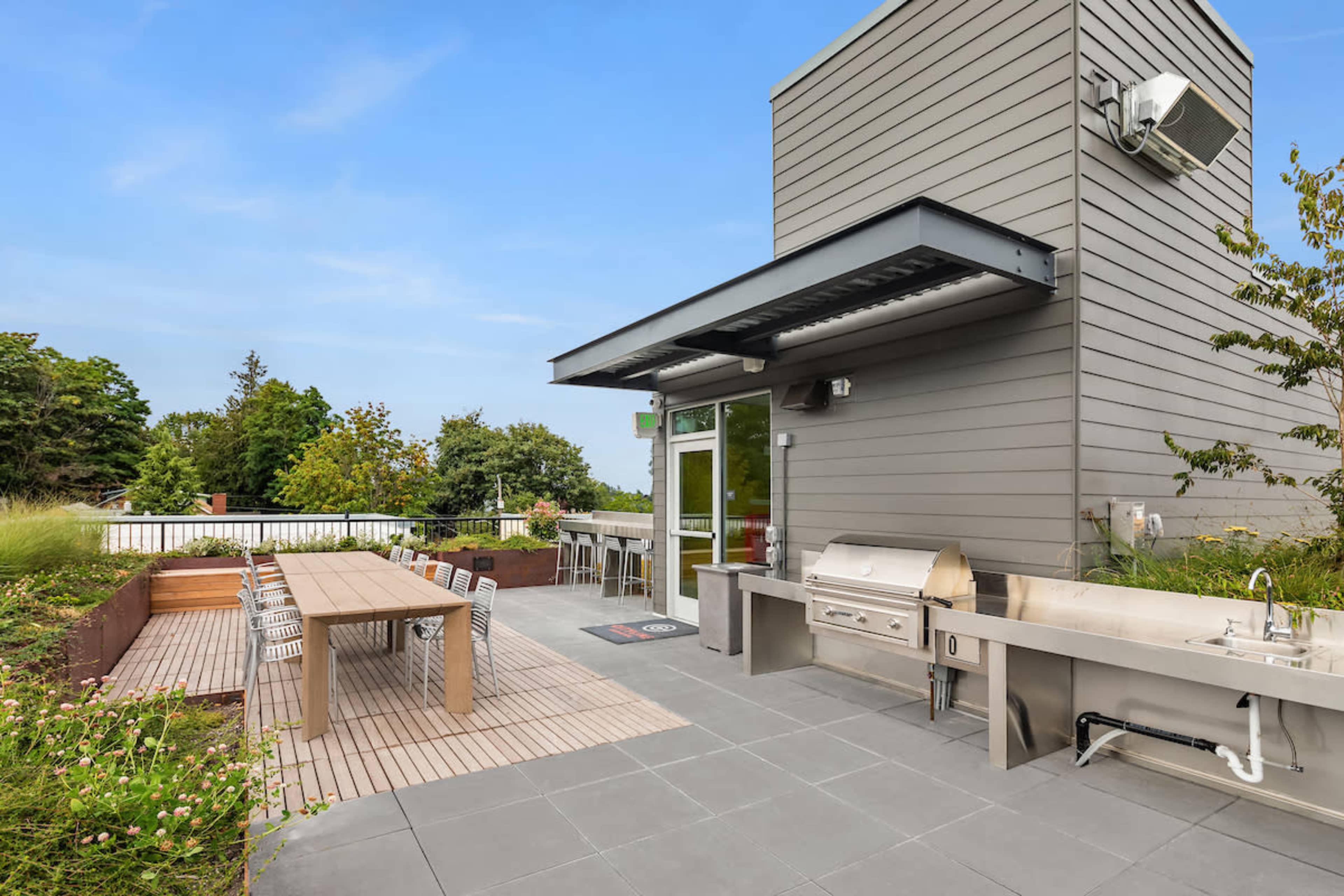 The image shows a modern rooftop patio featuring a dining table, a grill, and sleek outdoor furniture surrounded by greenery.