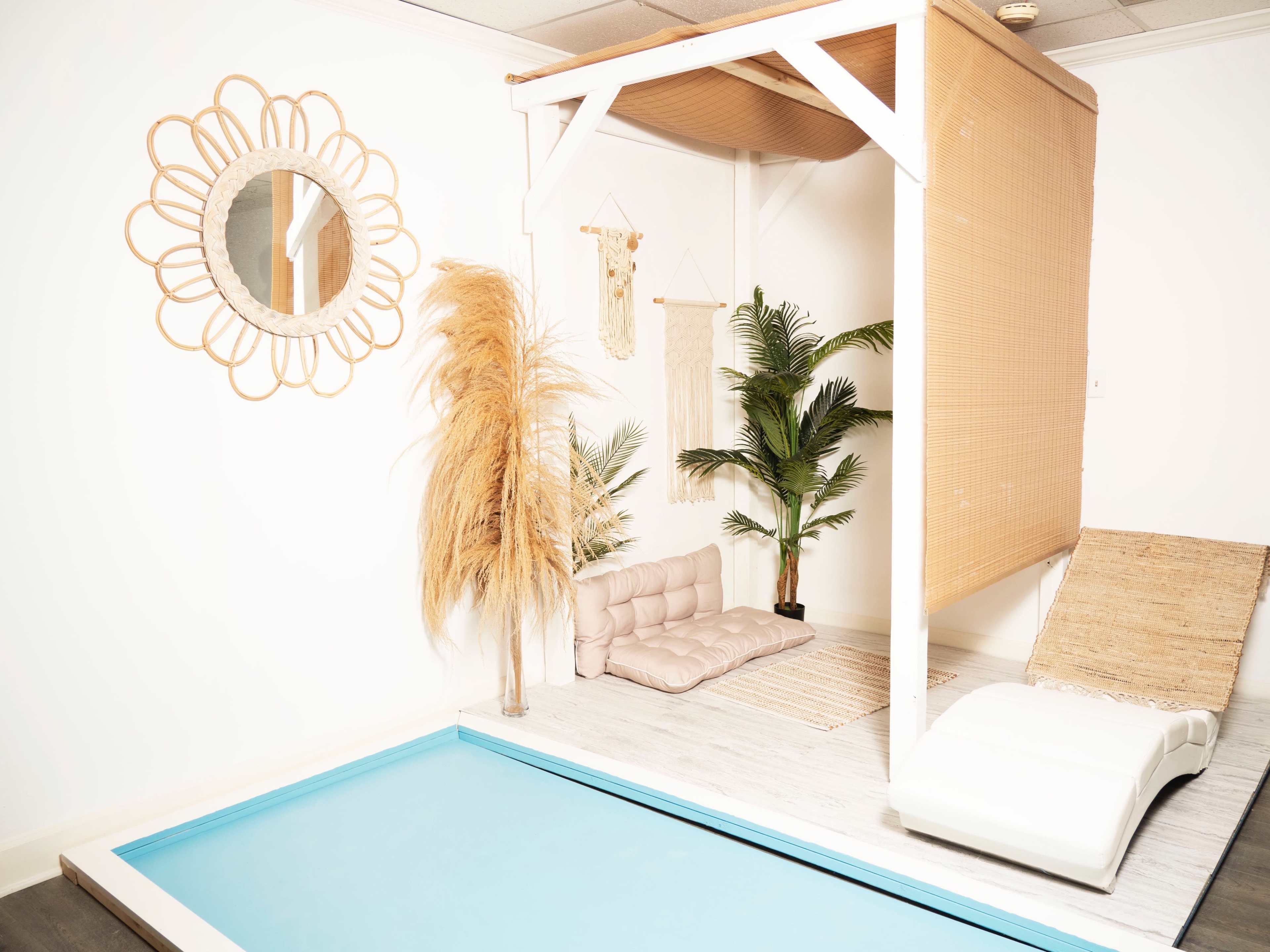 The image shows a relaxation area featuring a small pool, a raised platform with a cushion, indoor plants, and decorative wall hangings in a light-colored room.