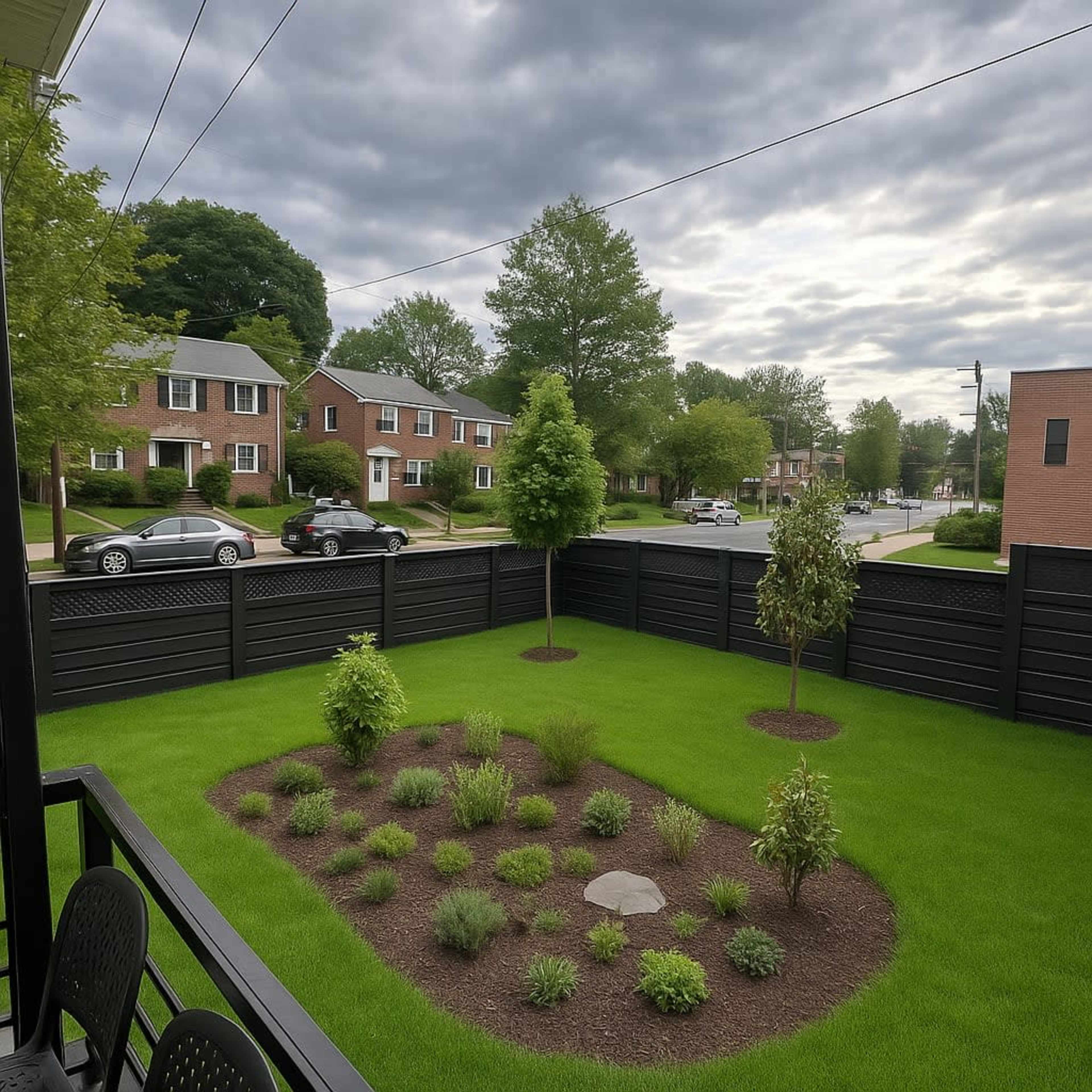 A well-maintained backyard features a small garden bed surrounded by grass and trees, enclosed by a wooden fence, with a view of residential streets and houses in the background.