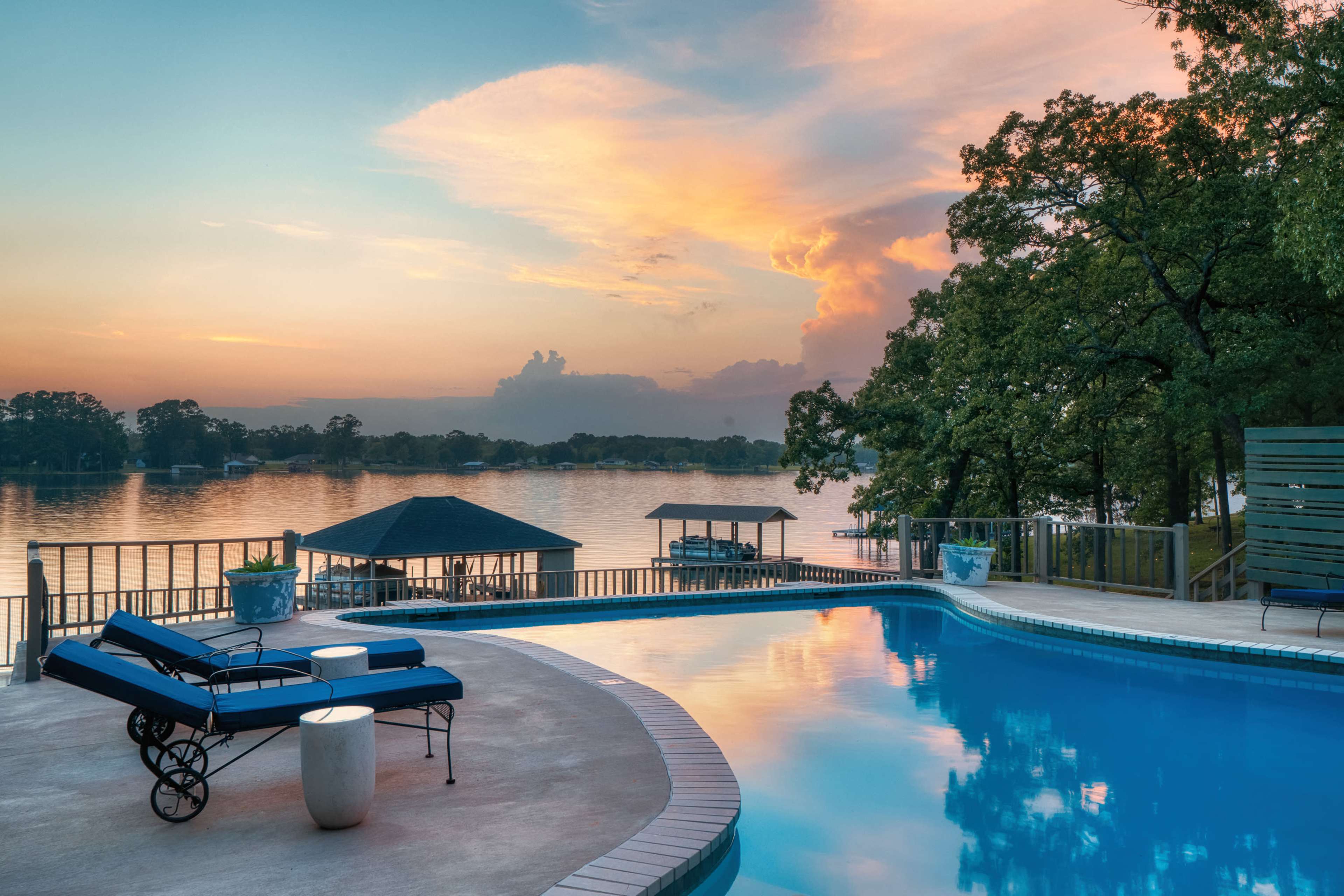 The image shows a swimming pool overlooking a calm lake at sunset, with lounge chairs positioned on the pool deck and trees in the background.