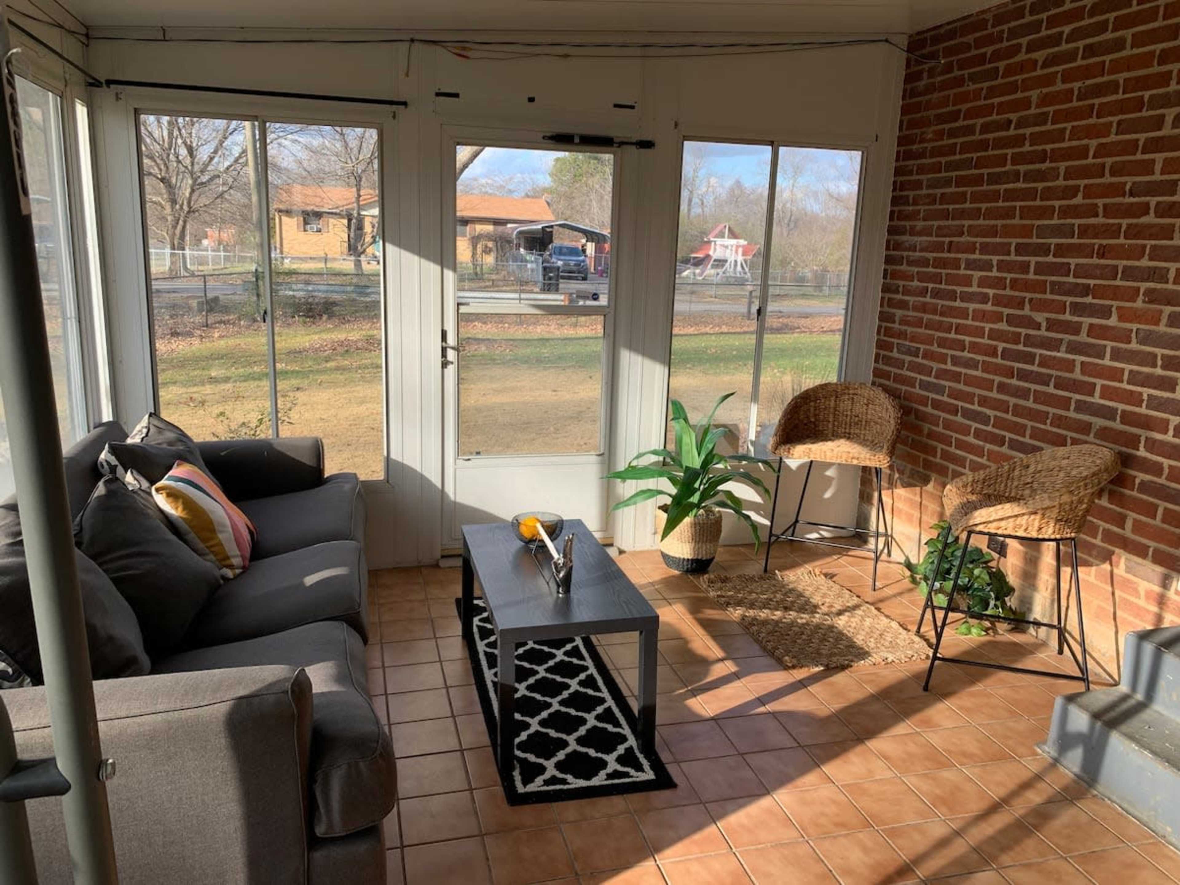 A sunlit sunroom with a gray sofa, a coffee table, and two wicker chairs, surrounded by large windows overlooking a grassy area and a road.