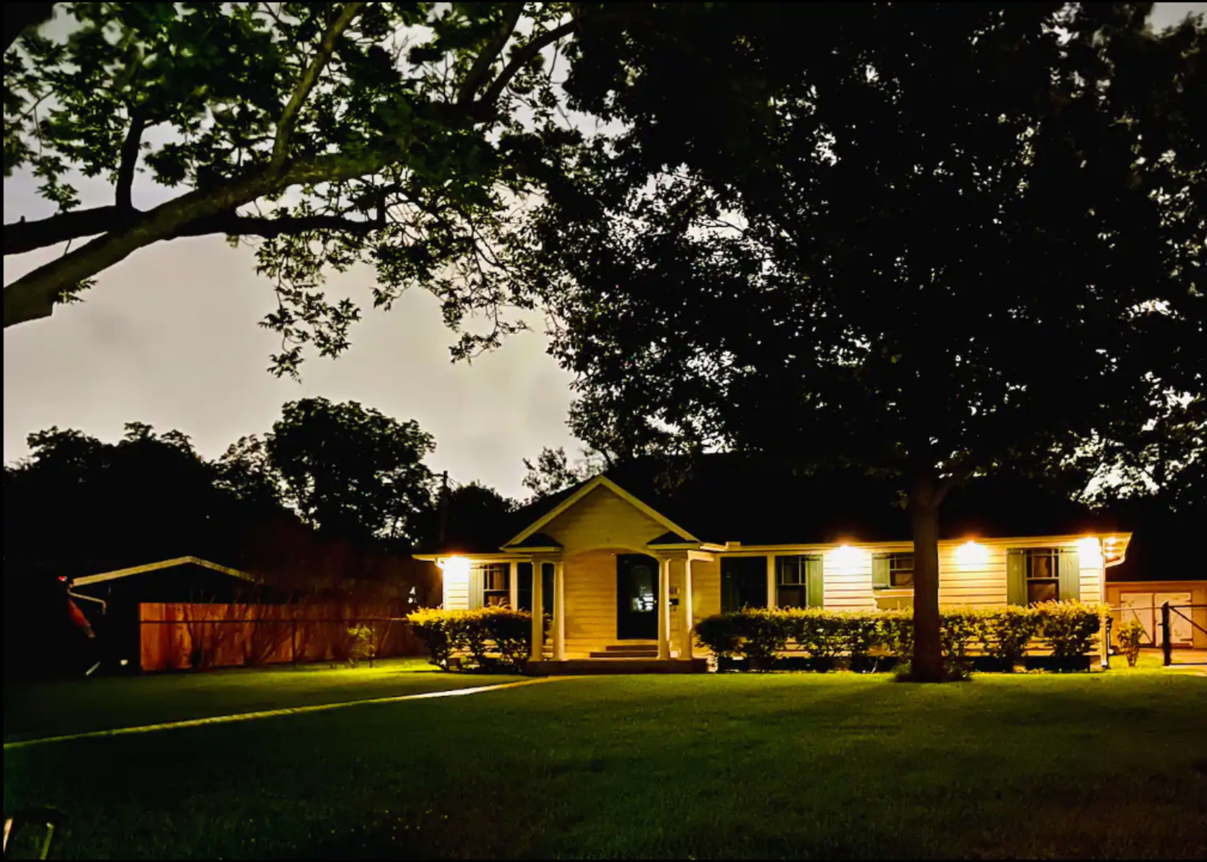 A single-story house with illuminated porch lights sits surrounded by trees in a dark, grassy yard at night.