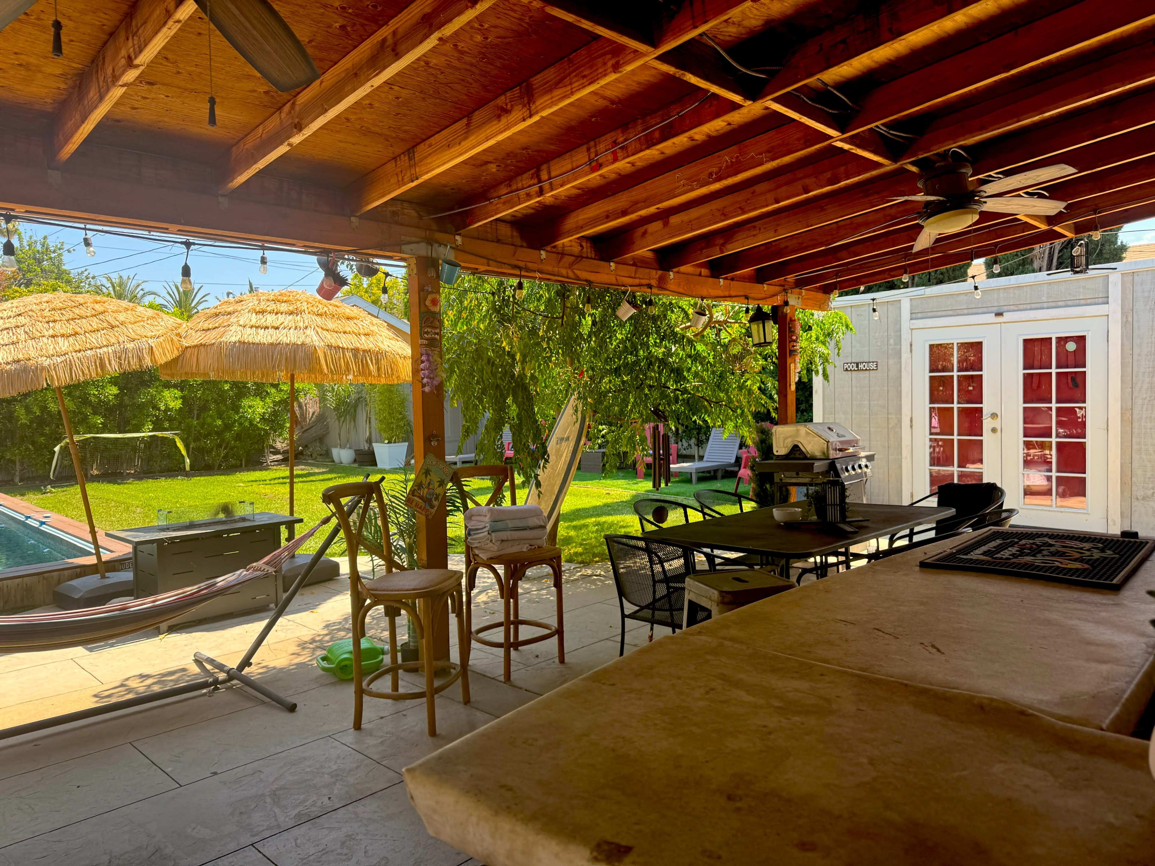 The image shows a covered outdoor area with a dining table, bar stools, and tropical-style umbrellas overlooking a green lawn and a pool.