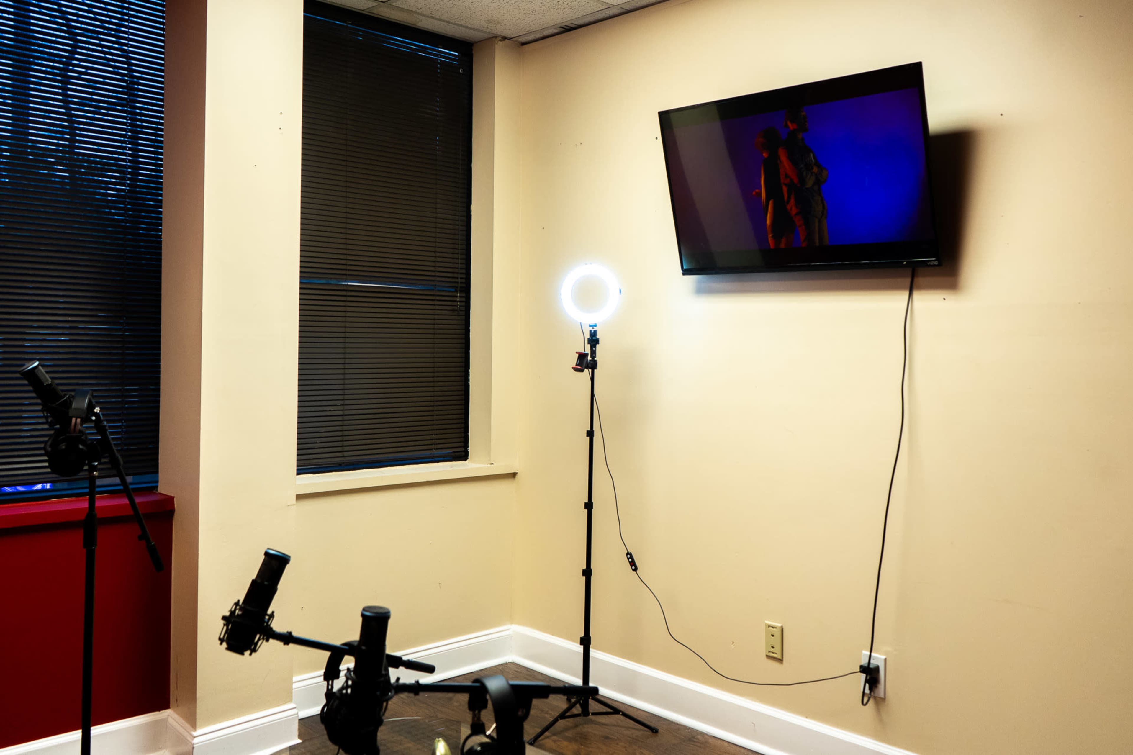 The image shows a small room with a television mounted on the wall, a ring light on a stand, and several microphones arranged on a table.