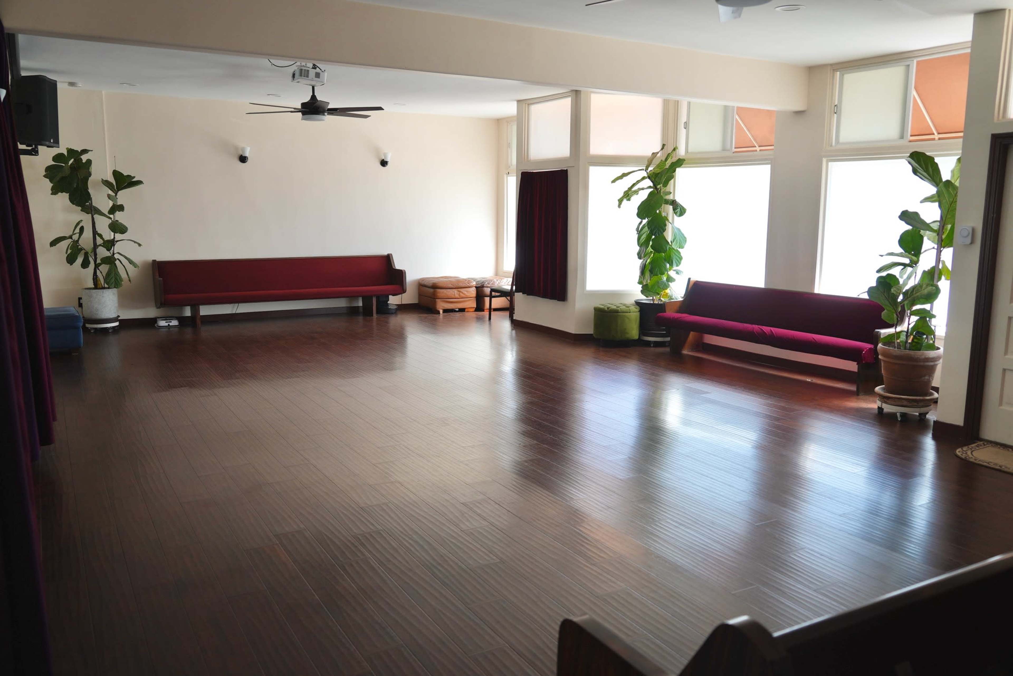 A spacious room with wooden flooring, two red benches against the walls, and several green plants near the windows.
