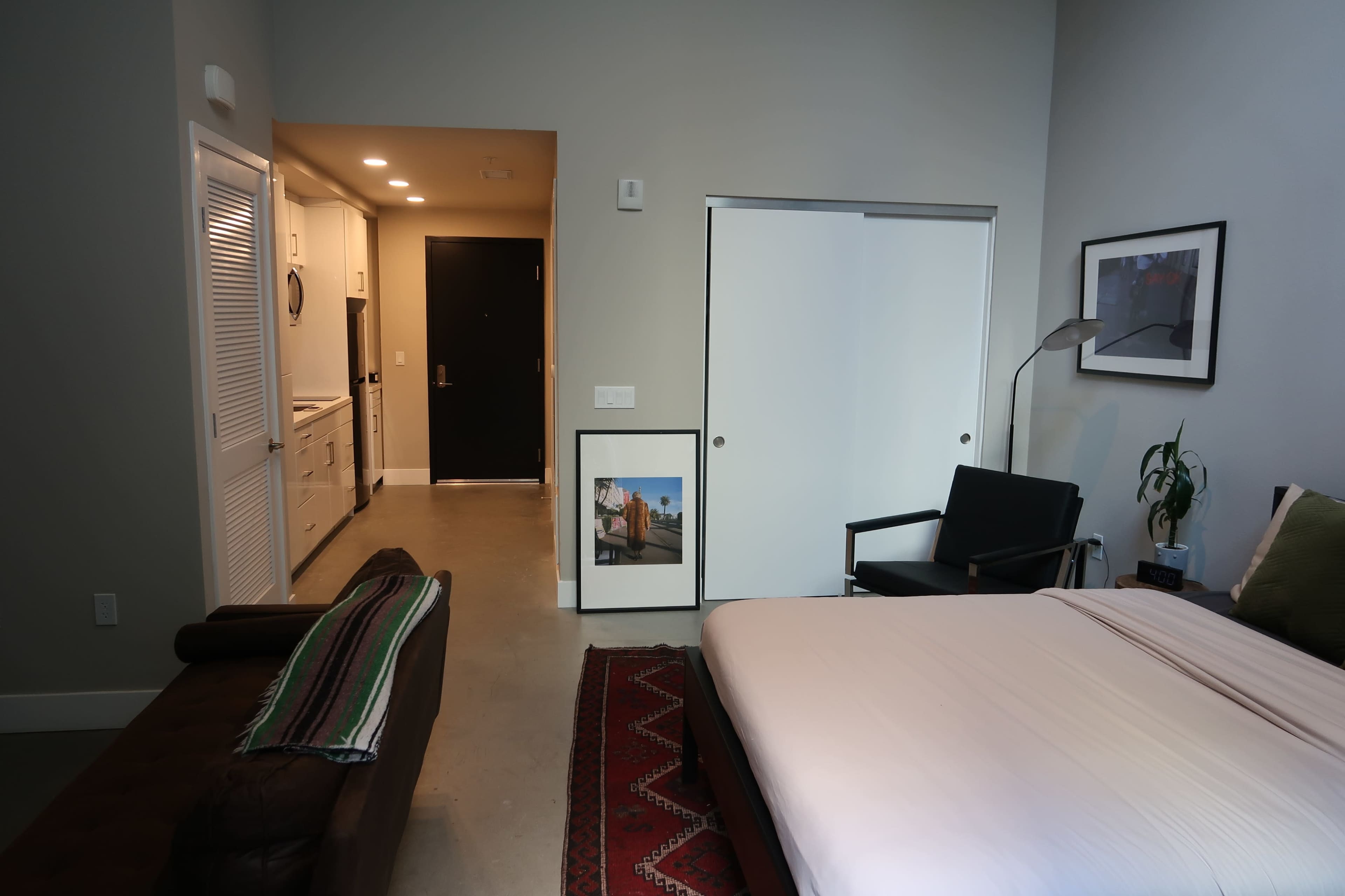 The image shows a minimalist bedroom featuring a bed with a light-colored sheet, a couch, a door leading to a kitchen area, and a framed photo on the wall.