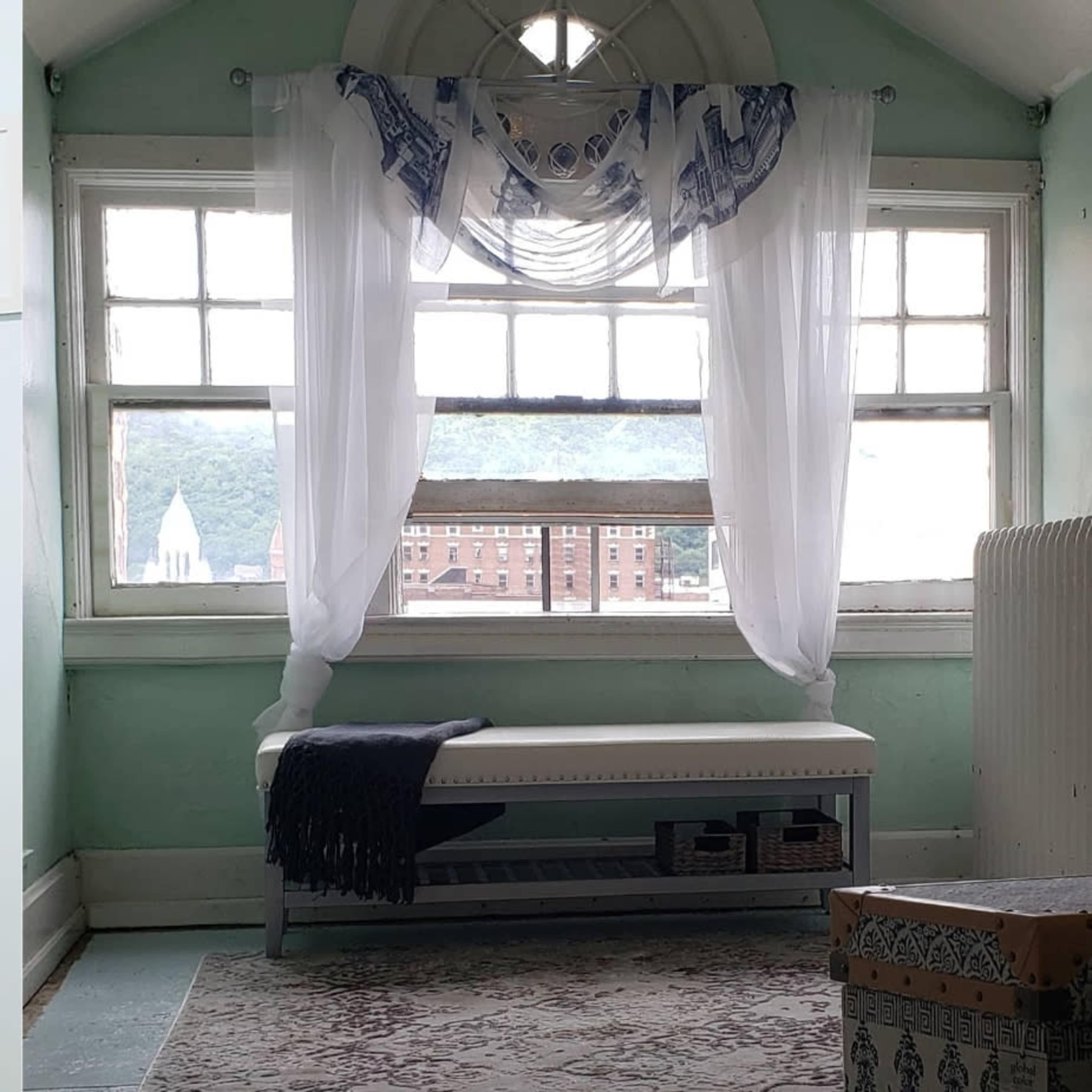 A sunlit room features a white window with sheer curtains, a bench with a blanket, and a view of buildings and hills outside.