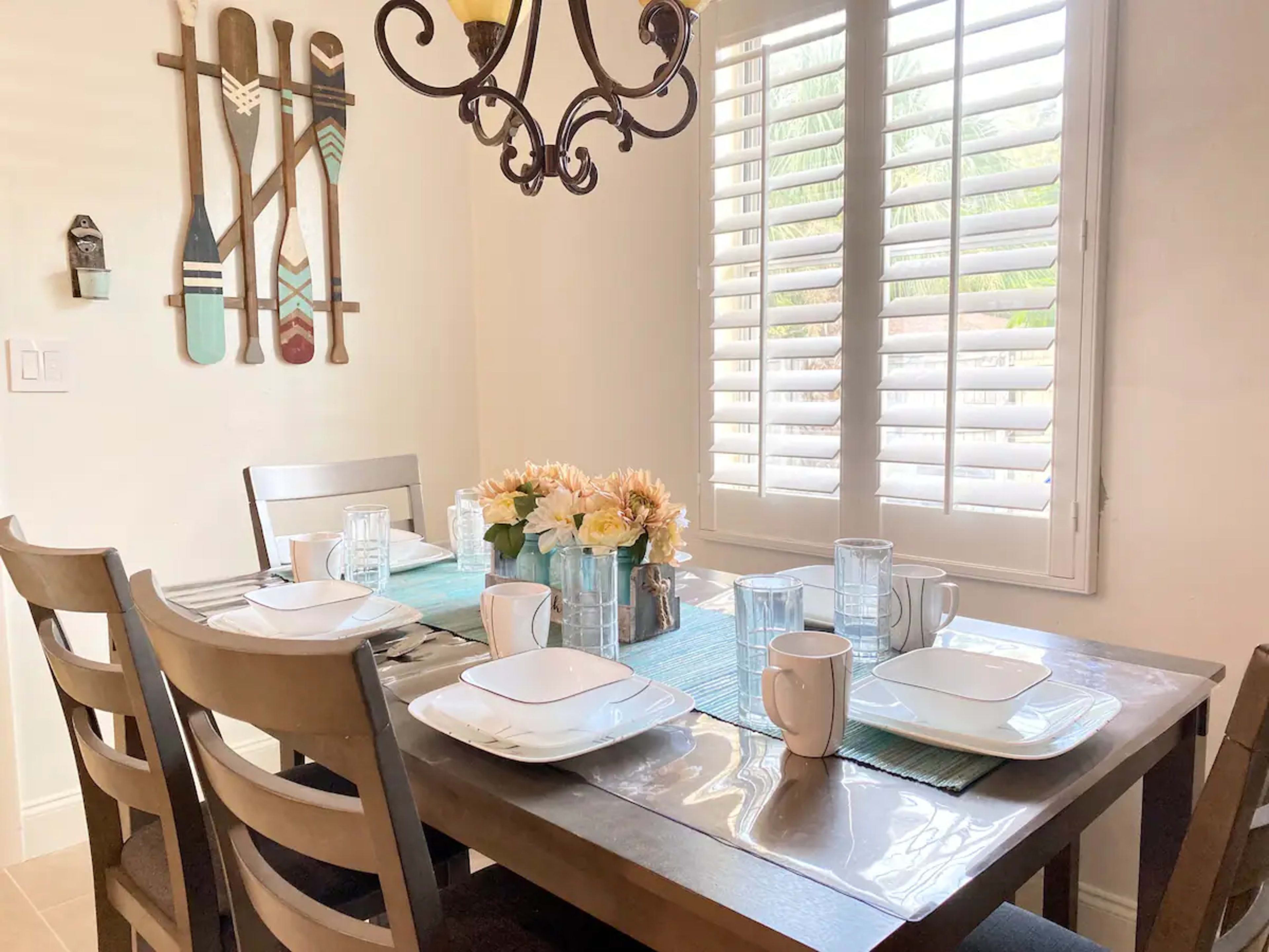 A dining table is set with plates, cups, and glasses, surrounded by chairs, with decorative paddles hanging on the wall and a flower arrangement in the center.
