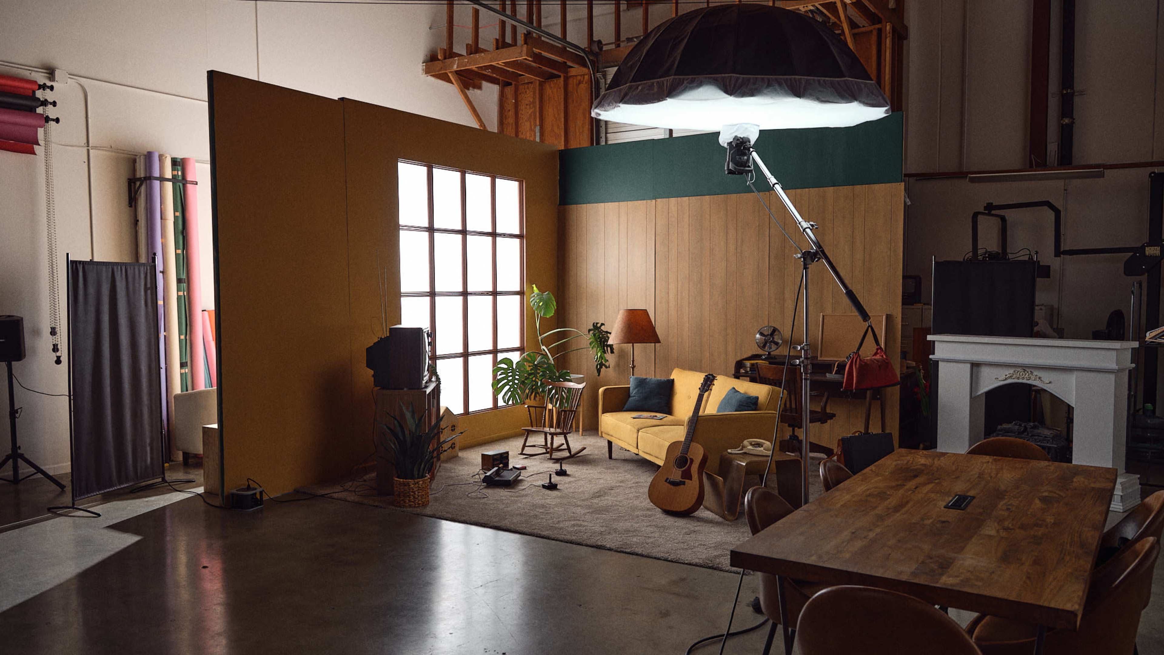 The image shows a well-lit studio space with a decorative backdrop, featuring a sofa, a guitar, plants, and various lighting equipment.
