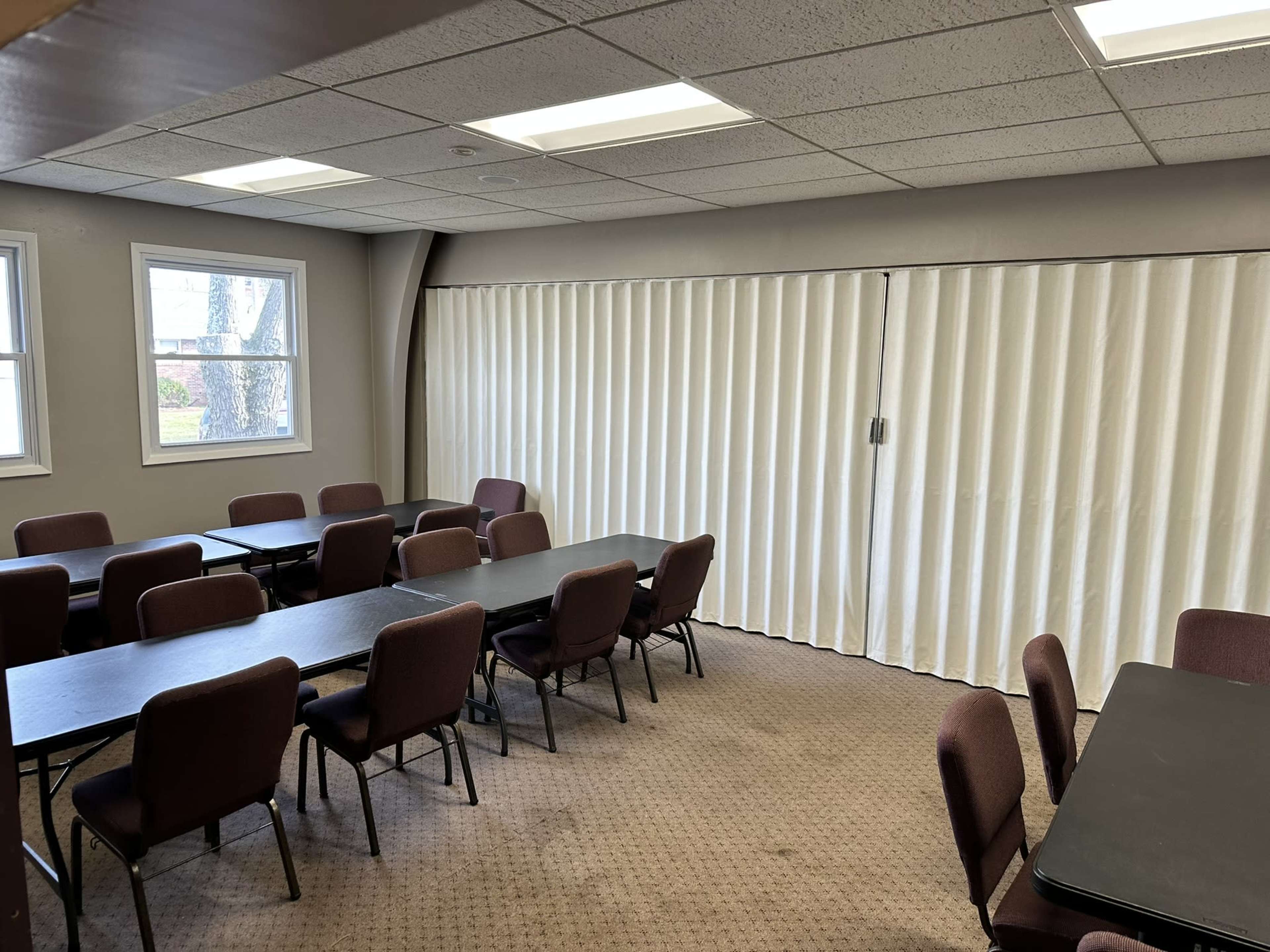 A meeting room with several tables arranged in rows and chairs along the sides, featuring a large curtain covering a window.