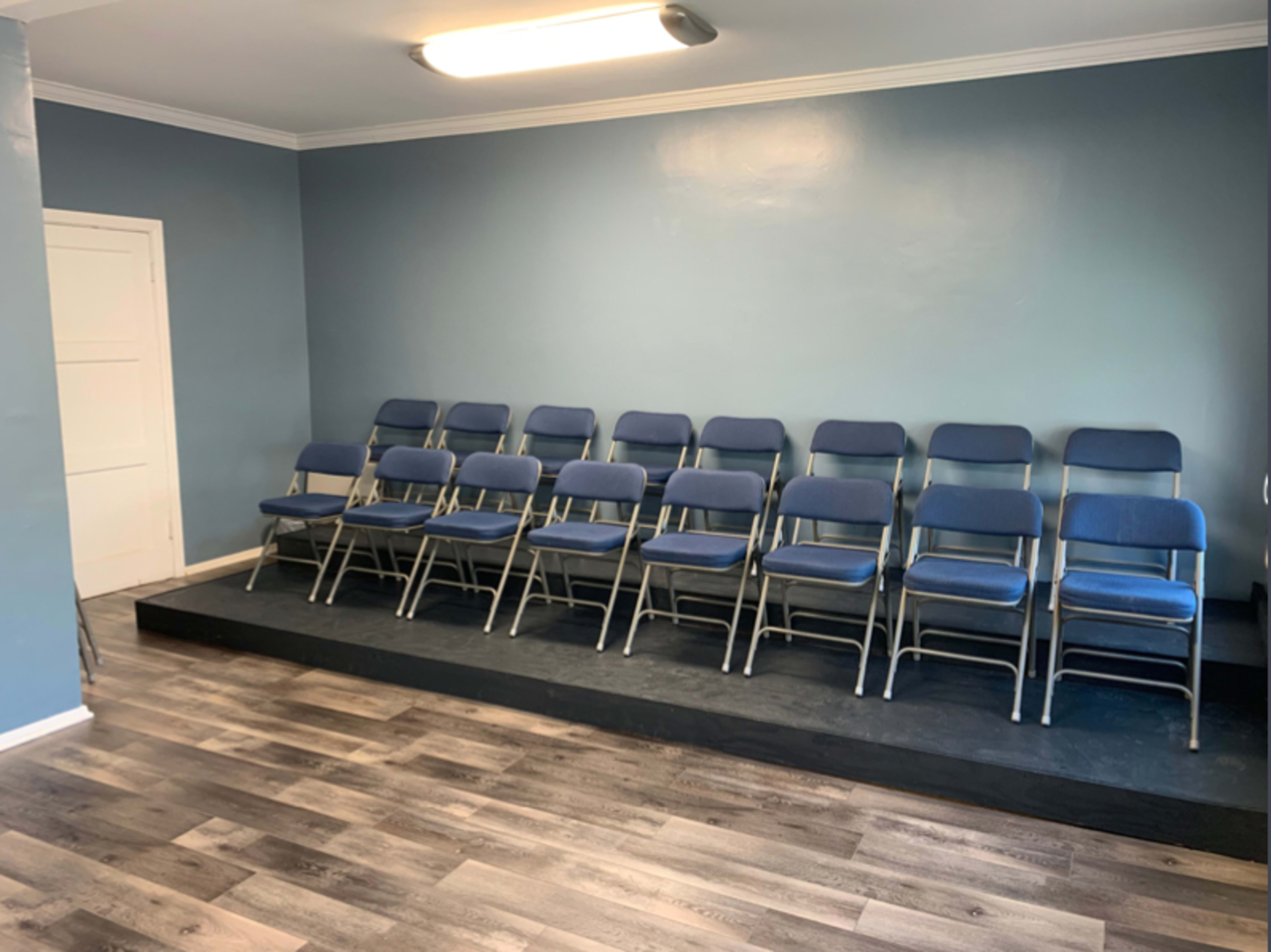 A row of blue folding chairs is arranged neatly on a raised platform in a sparsely decorated room with blue walls and a white door.