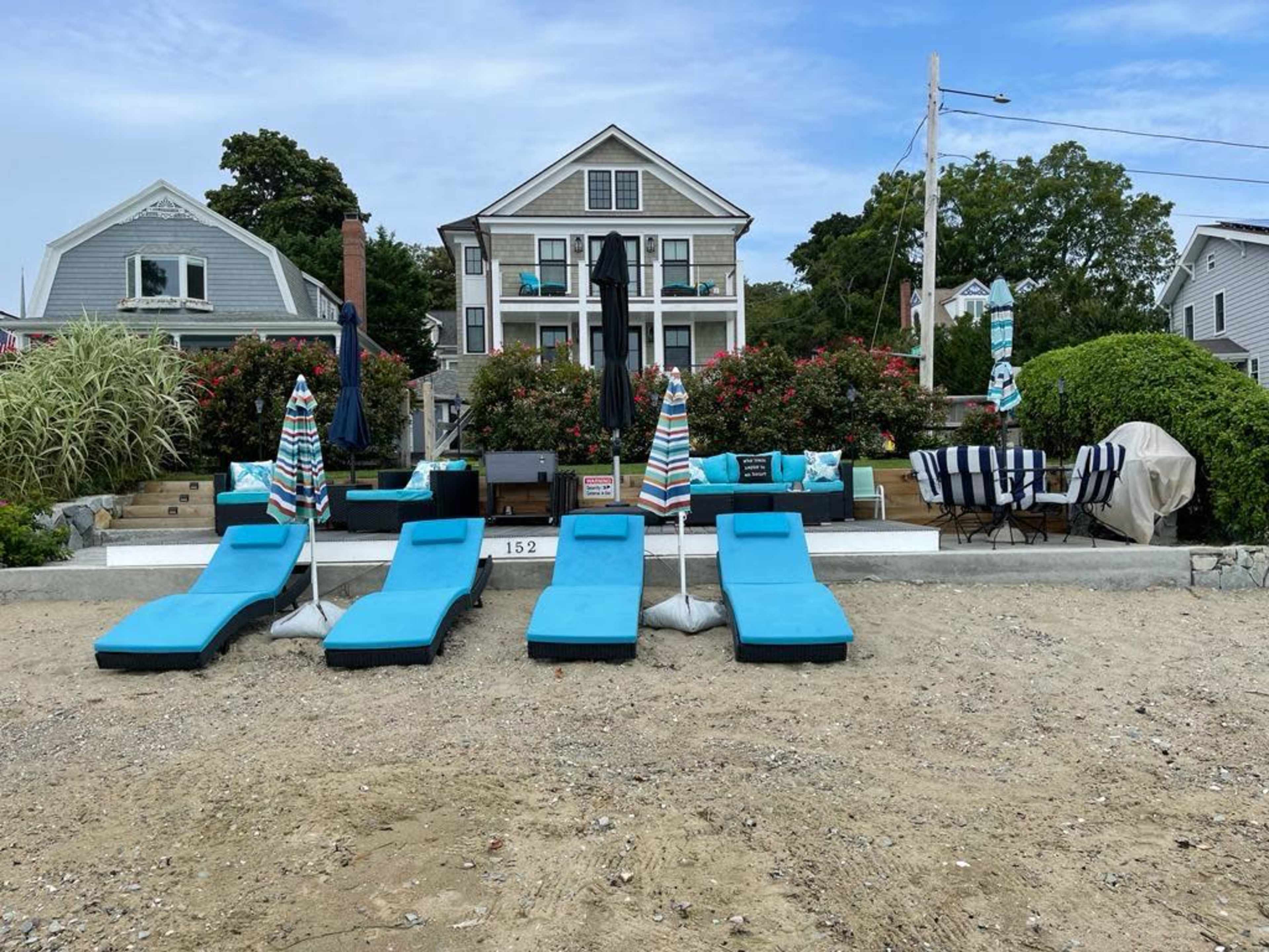 A beachside property featuring lounge chairs with umbrellas, a patio area, and a large house in the background surrounded by greenery.