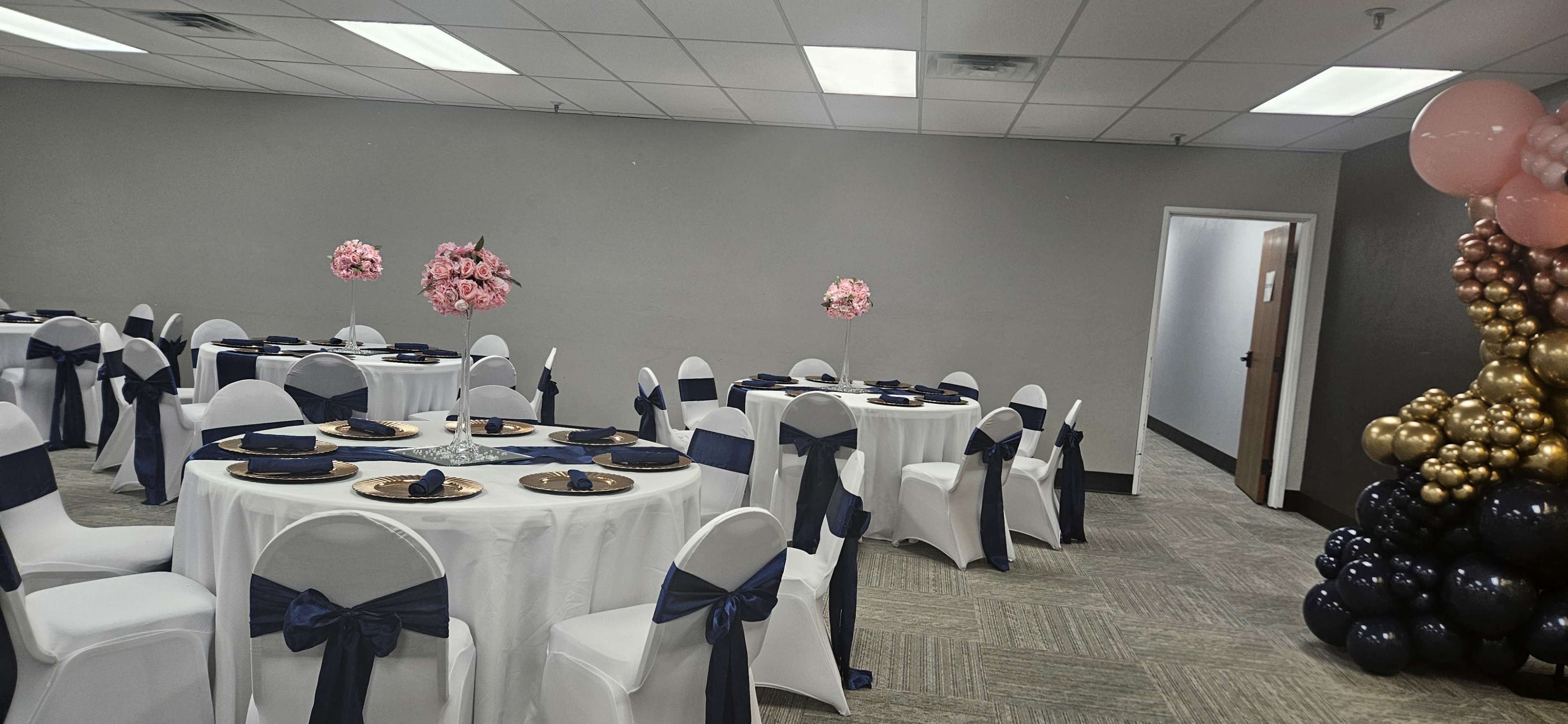 The image shows a banquet room set up with round tables covered in white tablecloths, navy blue ribbons on chairs, and decorative floral arrangements.