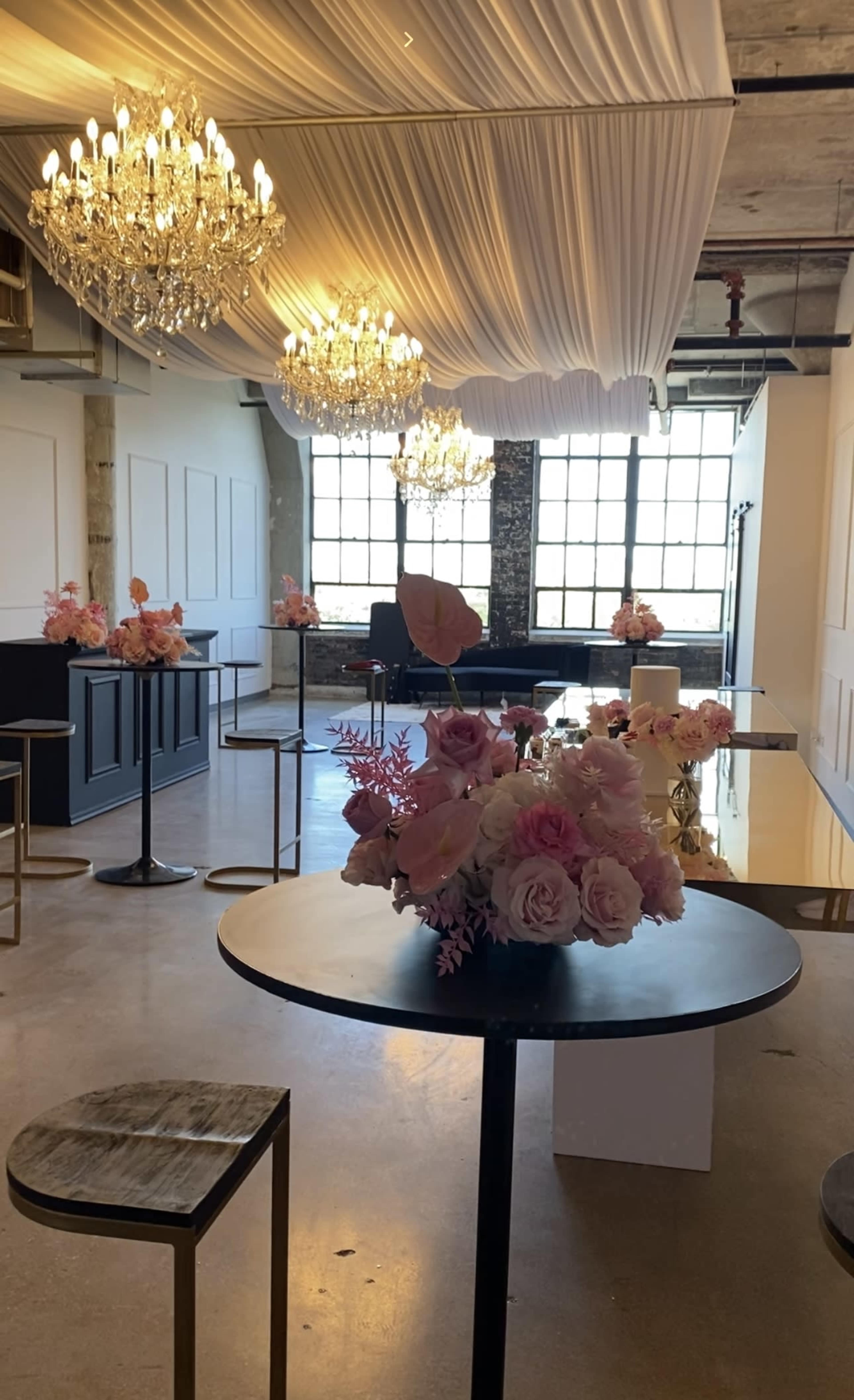 A spacious event venue with tables adorned with floral arrangements, illuminated by chandeliers hanging from a draped ceiling.