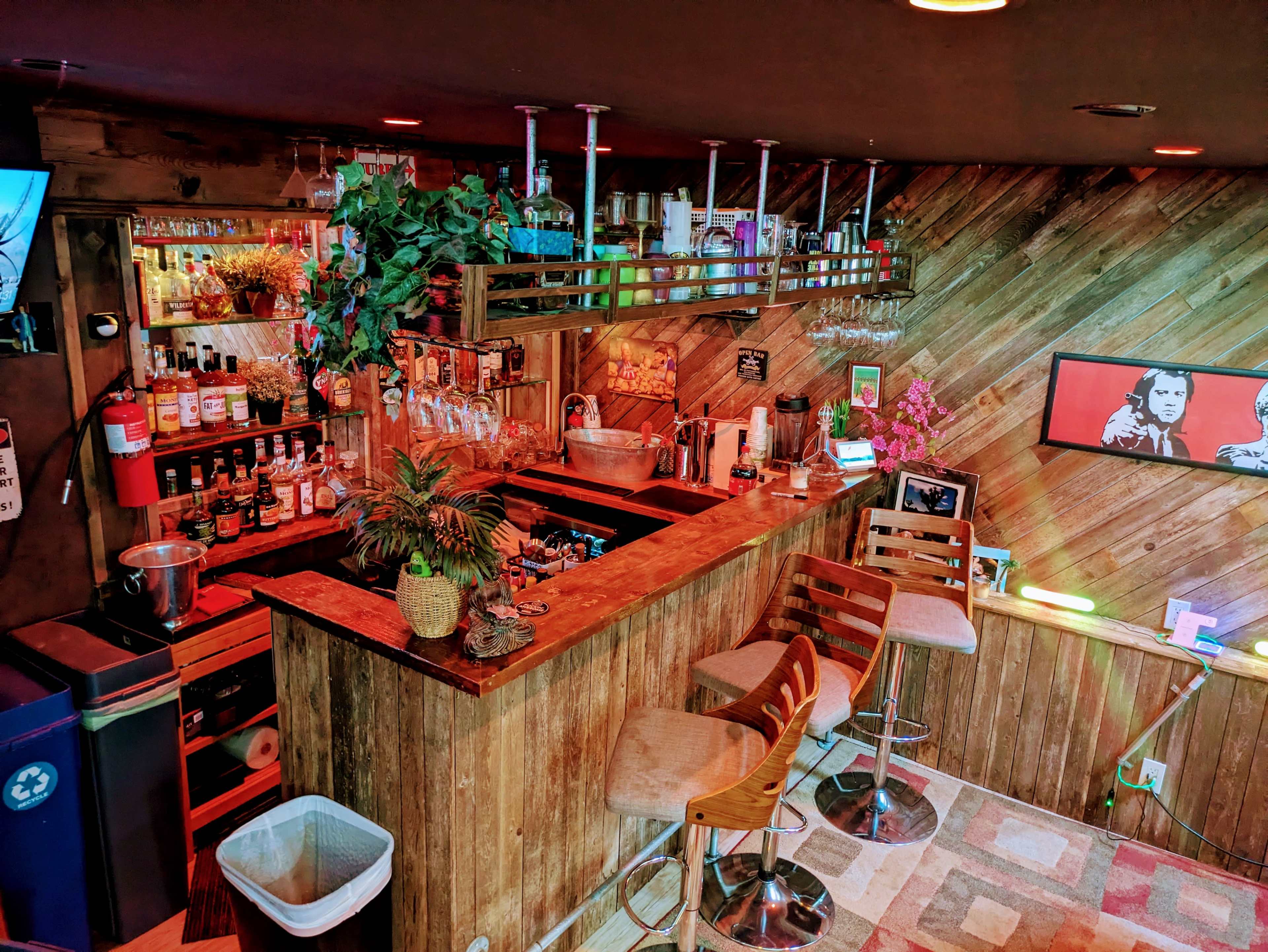 A home bar features wooden paneling, a variety of liquor bottles on shelves, and a seating area with high stools under soft lighting.
